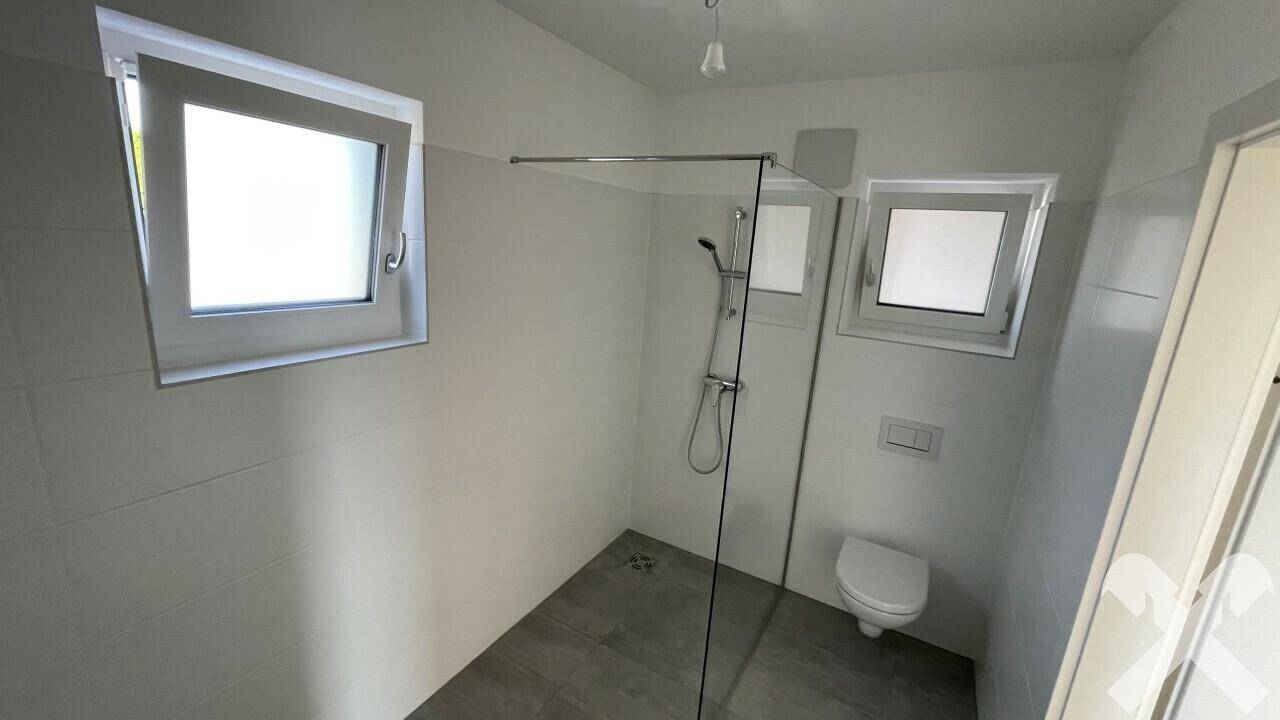 Helles Badezimmer mit begehbarer Dusche, wandhängender Toilette und zwei Fenstern.