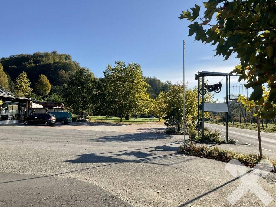 Großer asphaltierter Parkplatz neben einer Straße, umgeben von Bäumen und einem Motorrad-Schild.
