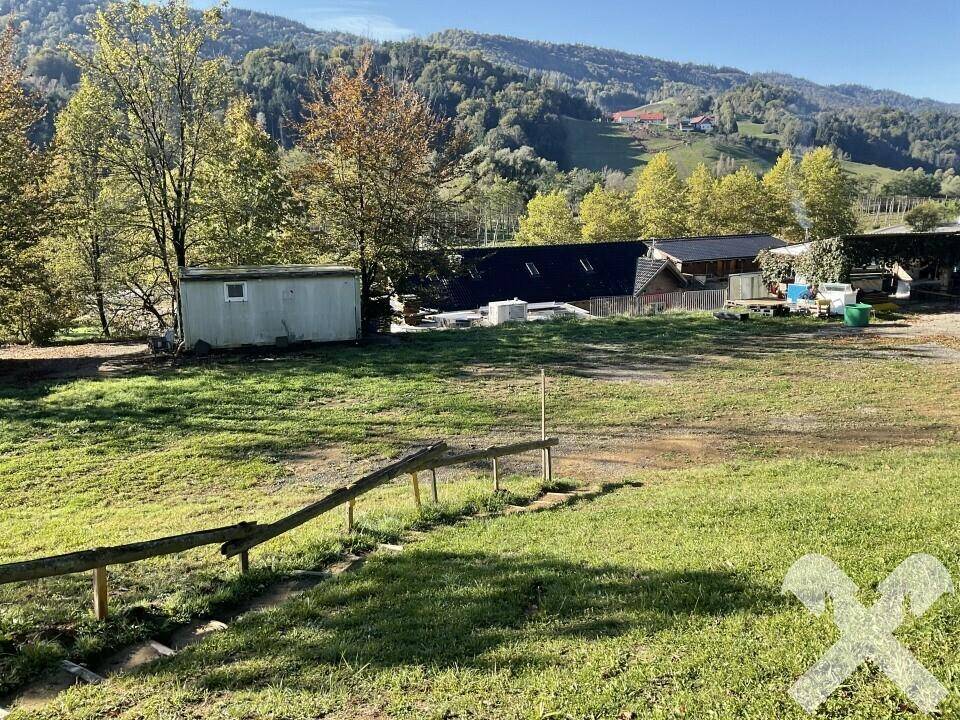 Offenes Gelände mit Gras, einem kleinen Schuppen und Blick auf die bewaldeten Hügel in der Ferne.
