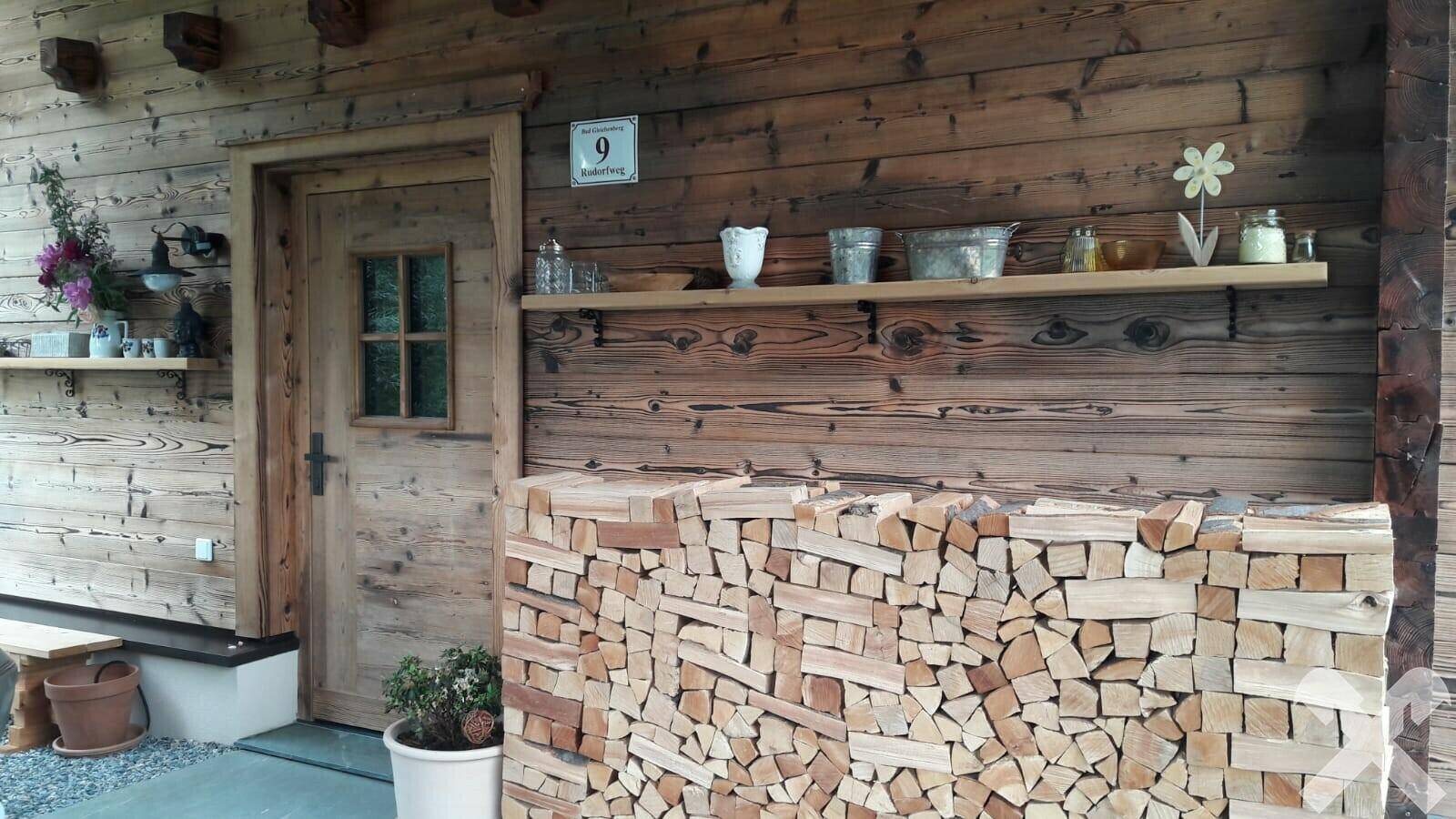 Eingangsbereich des Holzhauses mit gestapeltem Brennholz und rustikaler Holztür.