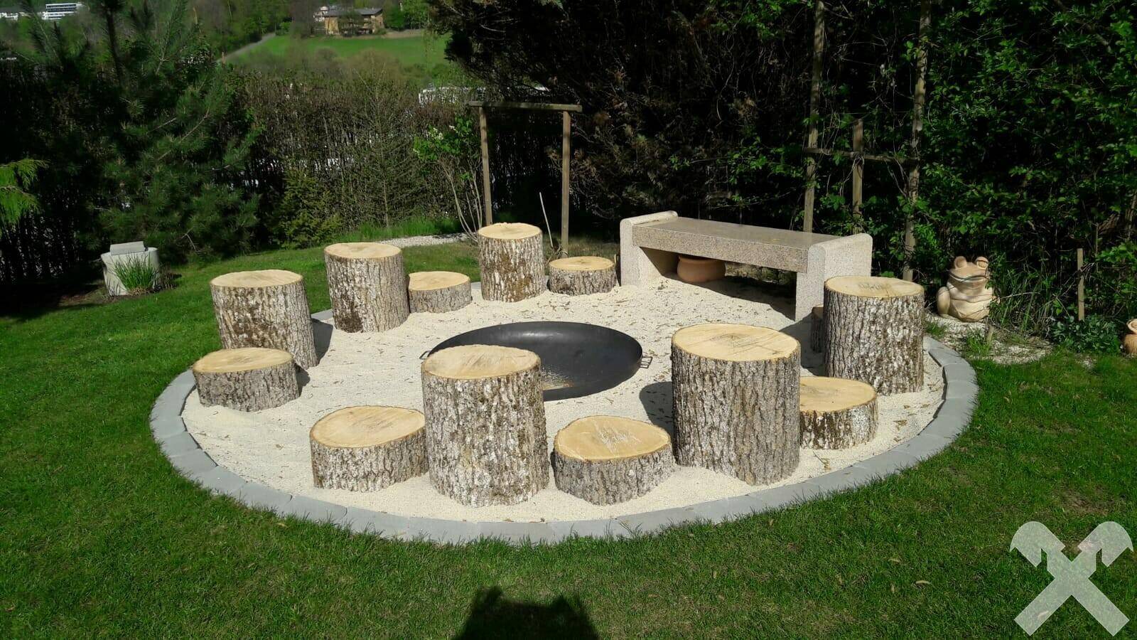 Gemütliche Feuerstelle im Garten mit Baumstumpf-Sitzgelegenheiten und Kiesuntergrund.
