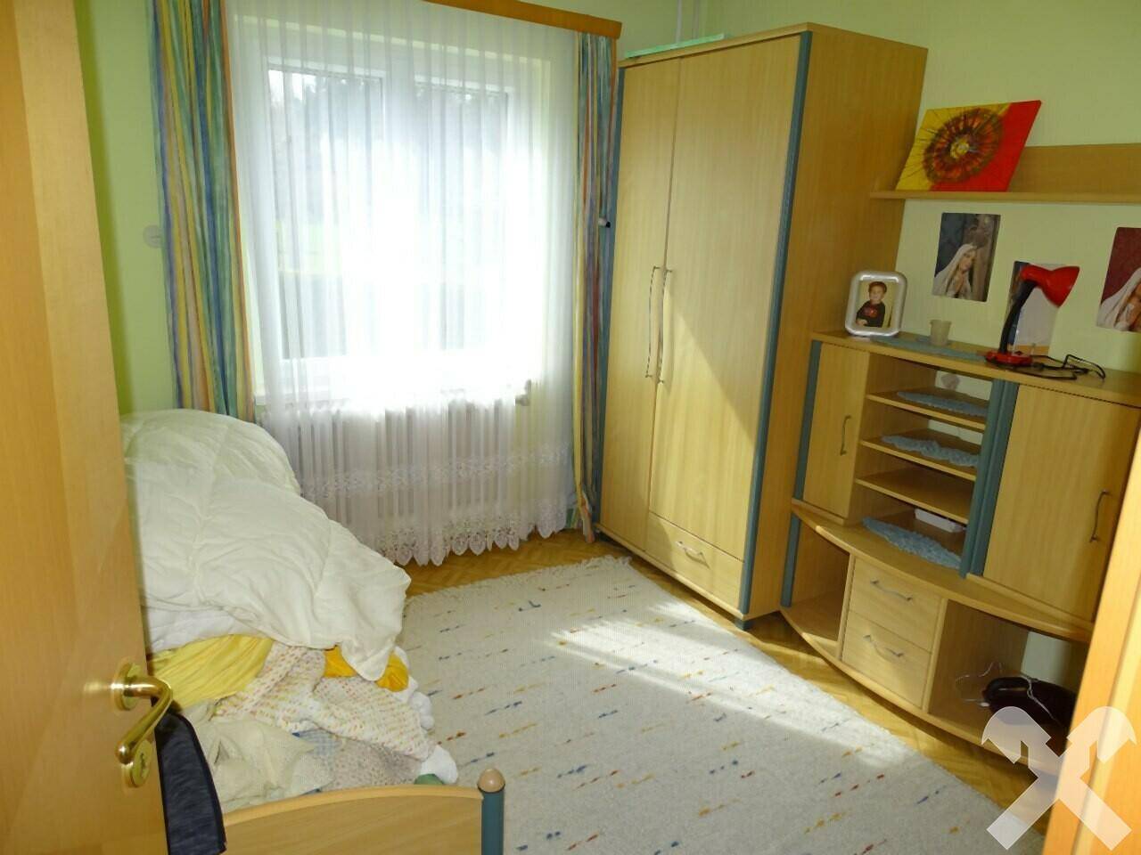 Schlafzimmer mit Einzelbett, Holzschrank und Fenster mit bunten Vorhängen.
