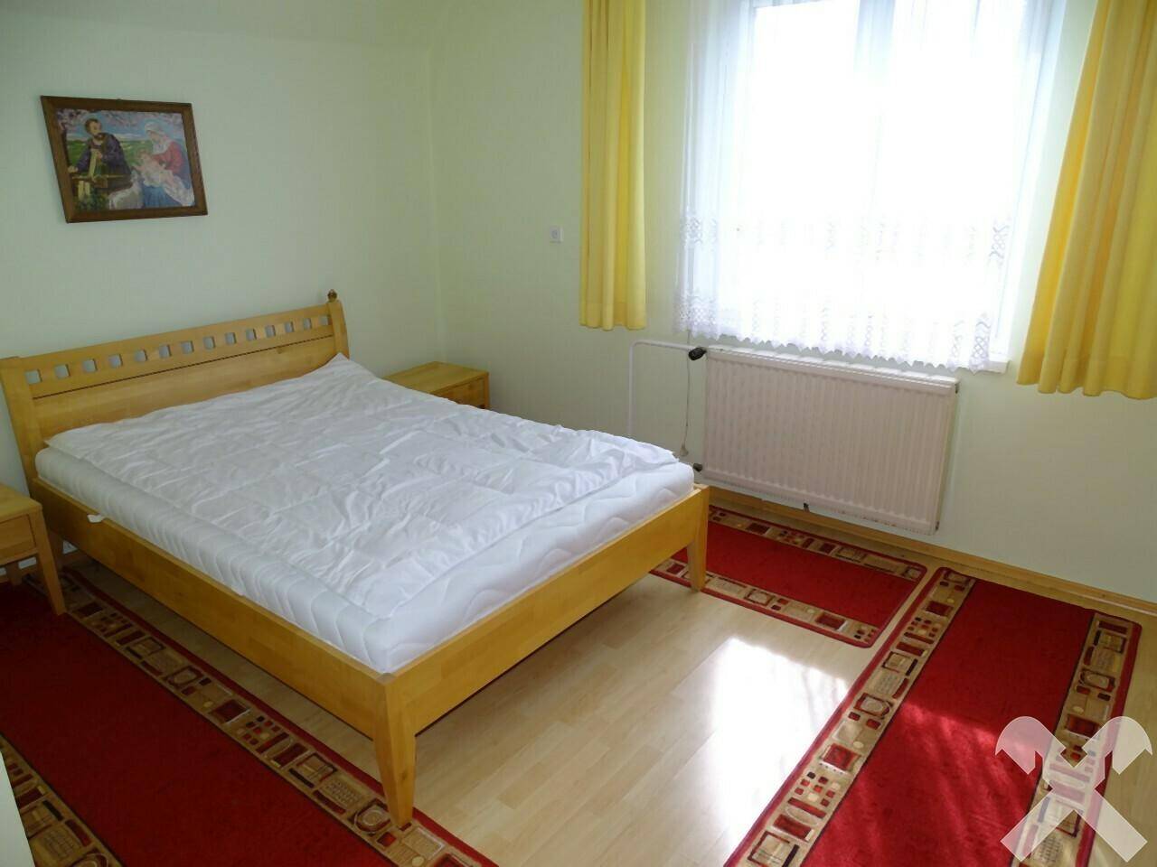 Schlafzimmer mit Holzdoppelbett, gelben Vorhängen und roten Teppichen auf Laminatboden.