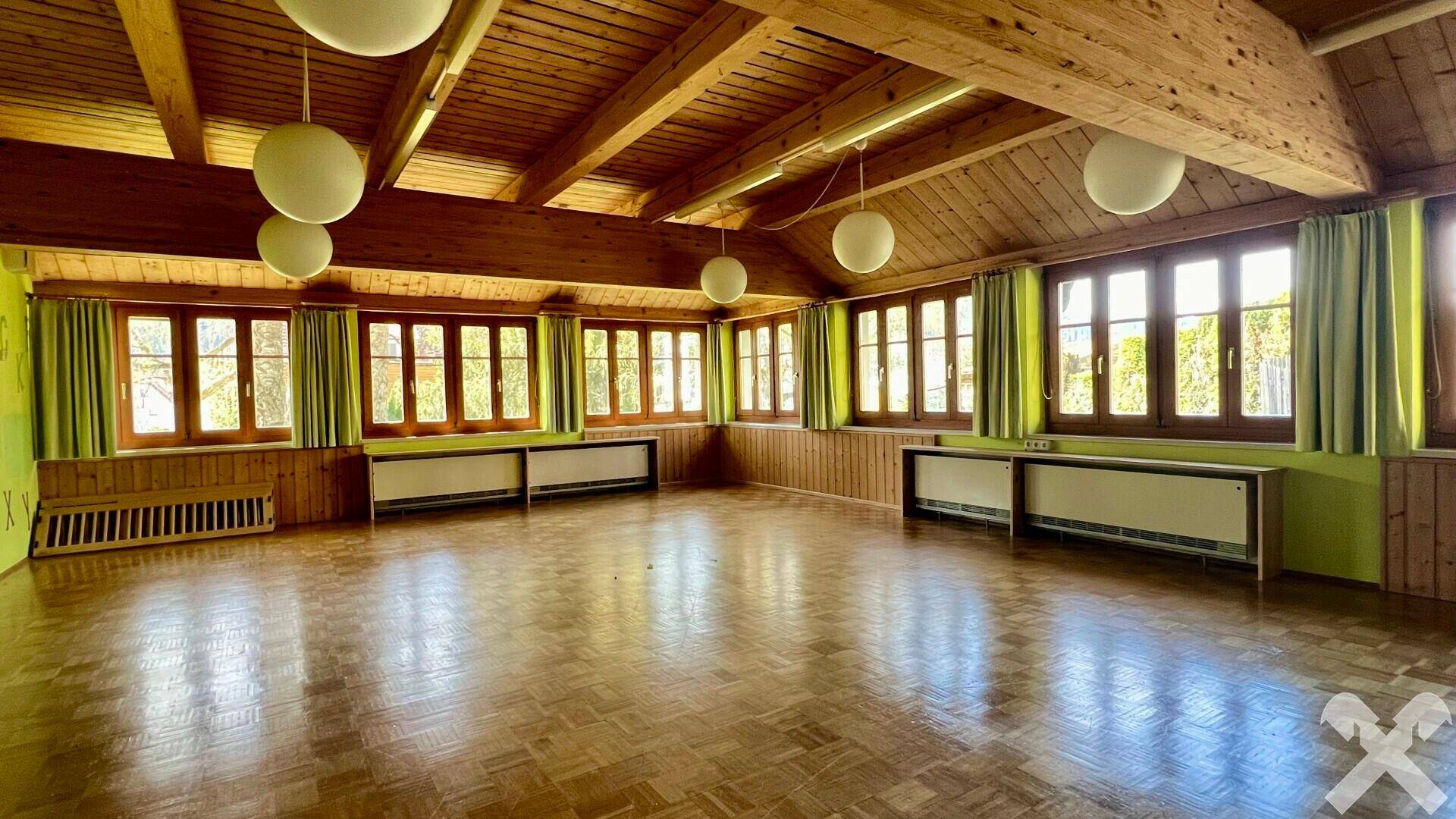 Großer heller Raum mit Holzbalkendecke, Parkettboden und vielen Fenstern mit grünen Vorhängen.