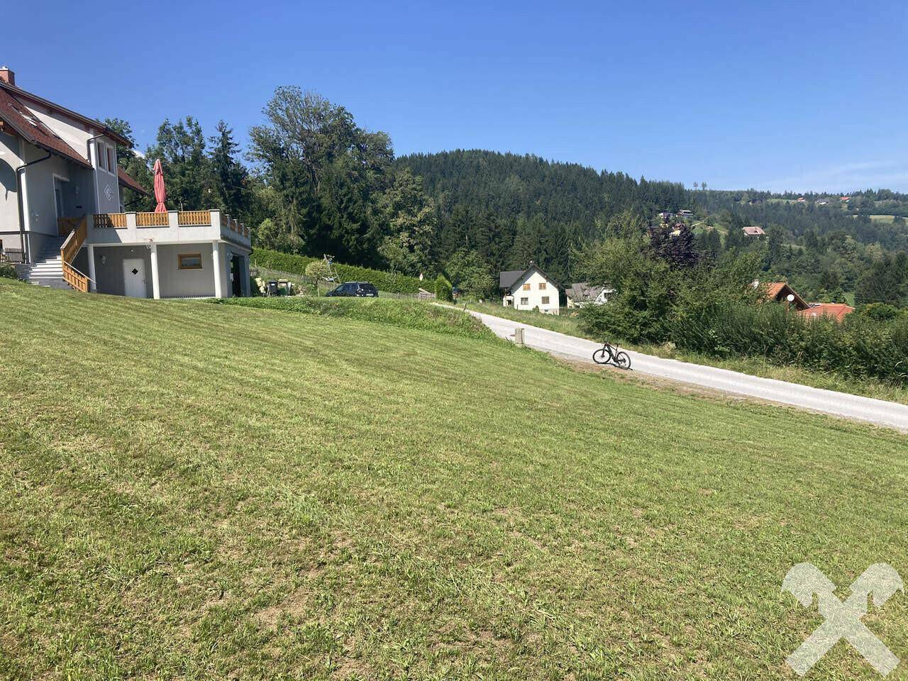 Blick auf ein Wohnhaus mit Balkon, eine Straße und bewaldete Hügel in einer ländlichen Umgebung.