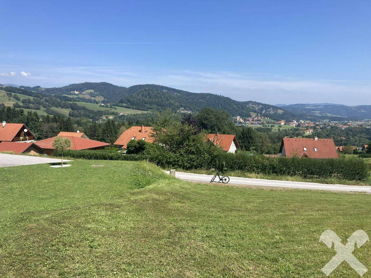 Panoramablick über eine grüne Wiese mit Häusern und bewaldeten Hügeln am Horizont unter blauem Himmel.