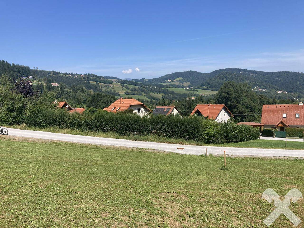 Blick über eine grüne Wiese auf eine Reihe von Häusern mit roten Dächern und bewaldeten Hügeln.