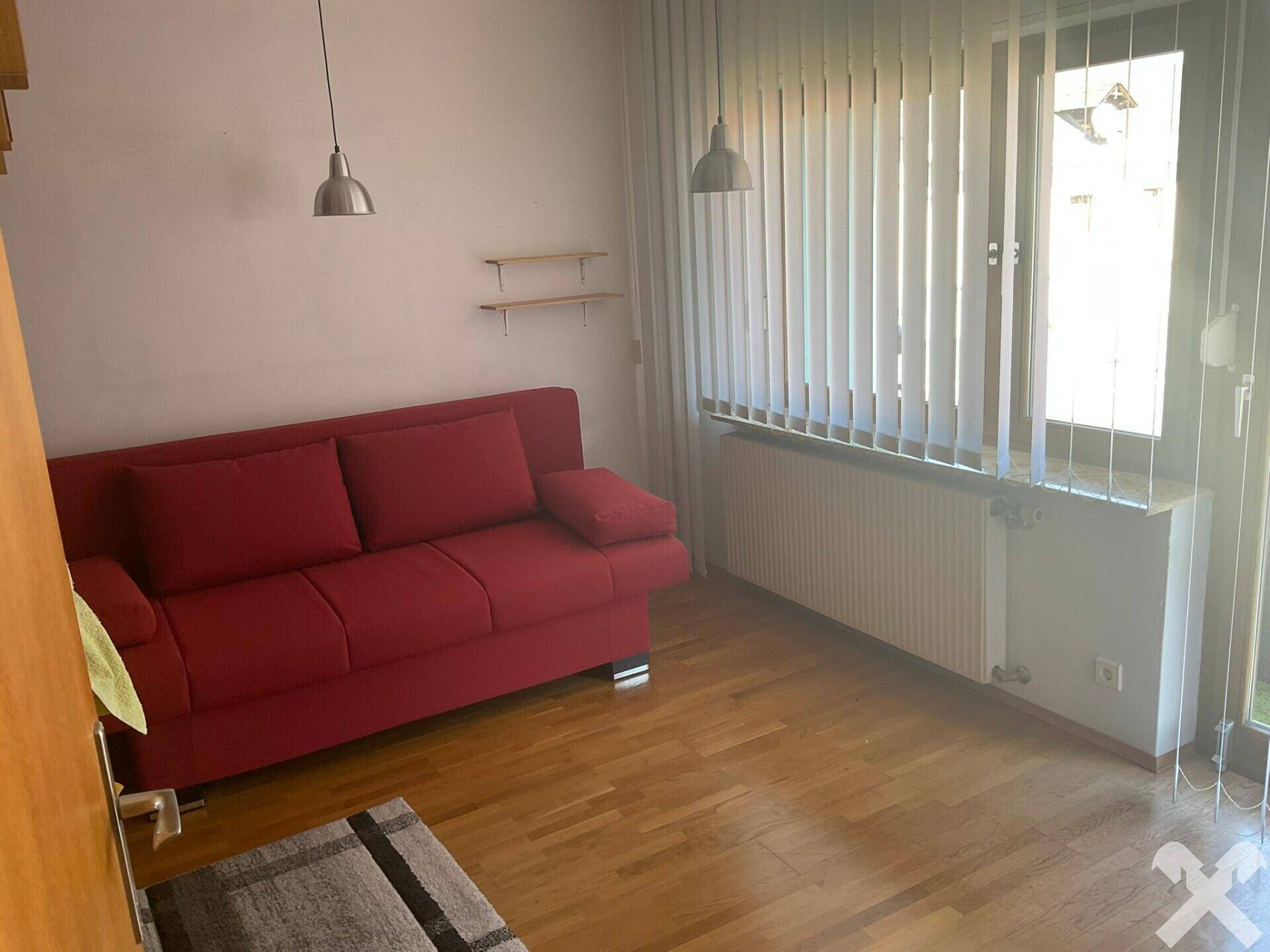 Helles Zimmer mit rotem Sofa, Holzfußboden und vertikalen Jalousien.