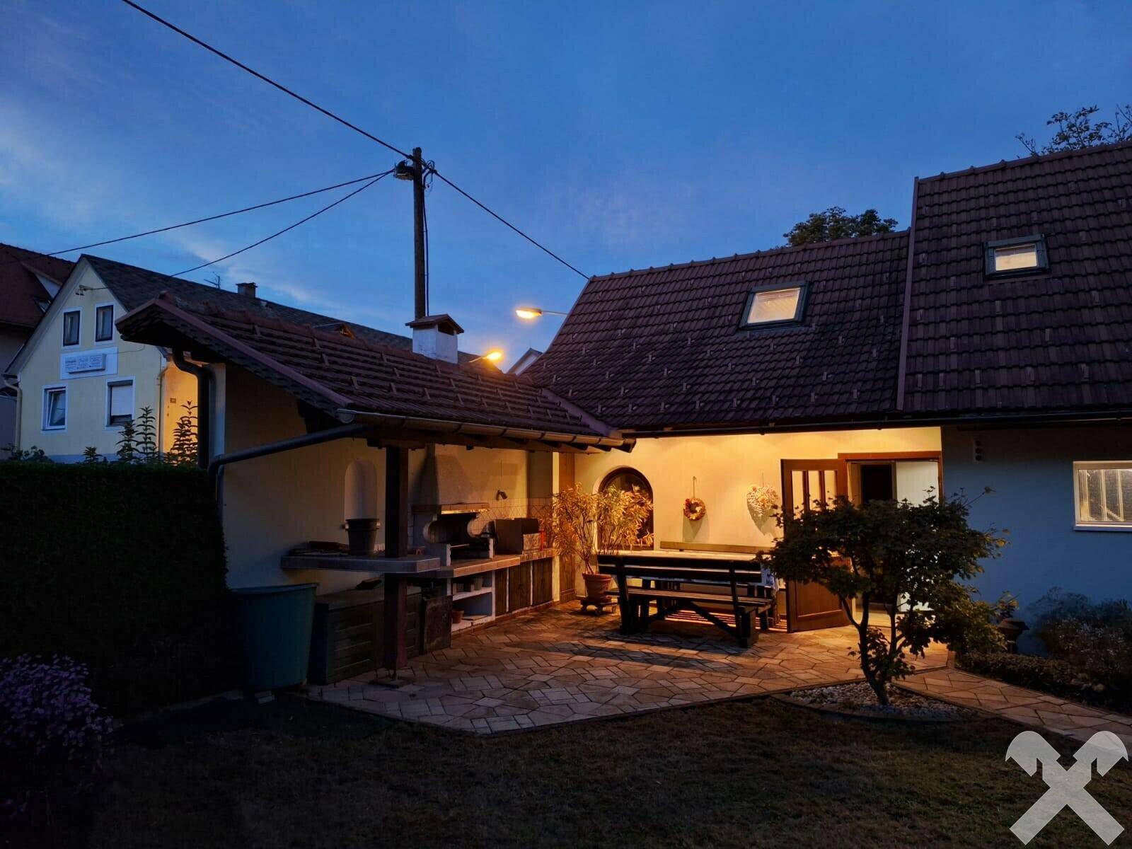 Abendliche Ansicht des Außenbereichs mit überdachtem Grillplatz und Sitzgelegenheiten.