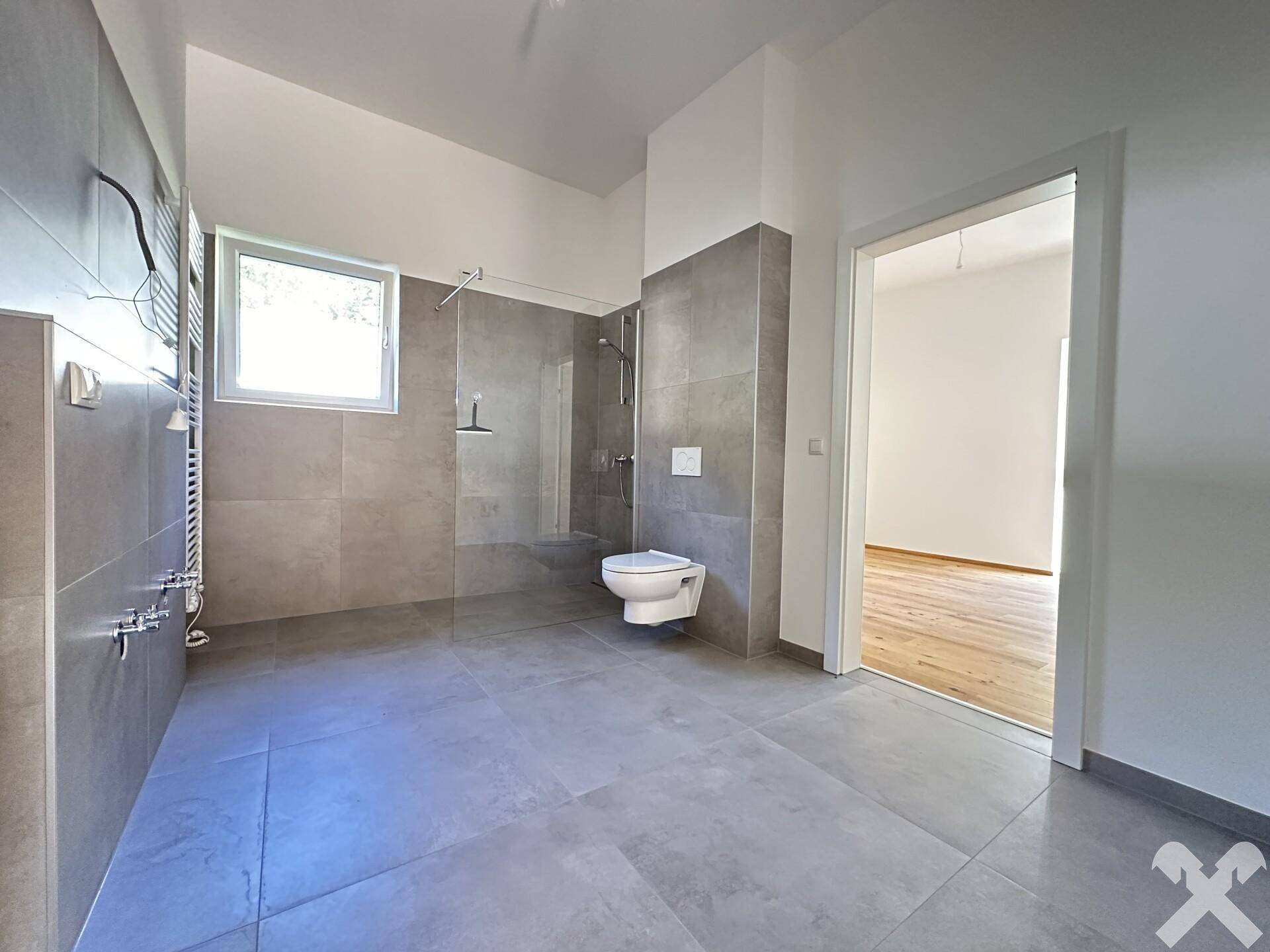 Modernes Badezimmer mit großer begehbarer Dusche, wandhängendem WC, Fenster und grauen Fliesen.