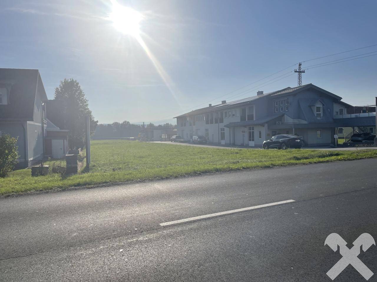 Weitläufiges grünes Grundstück an einer Straße, umgeben von Gebäuden unter strahlendem Sonnenschein.