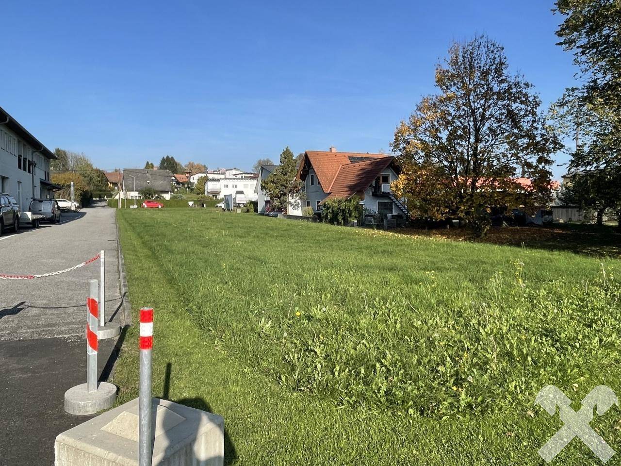 Unbebautes Grundstück mit hohem Gras entlang einer Straße, umgeben von Wohnhäusern und Bäumen.