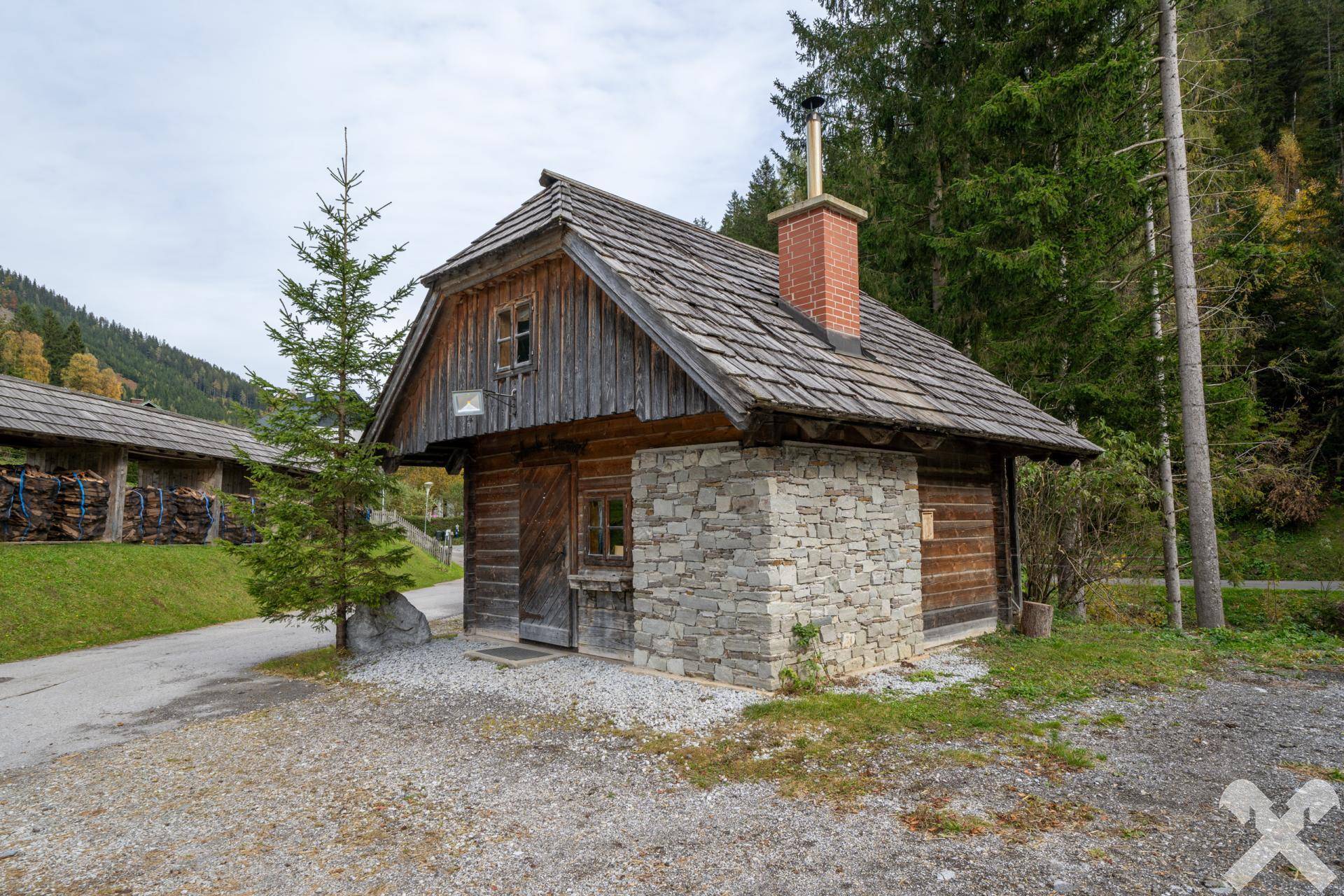 Kleines, rustikales Nebengebäude mit Holzschindeldach und Steinmauerwerk, umgeben von Bäumen und einer Schotterfläche.