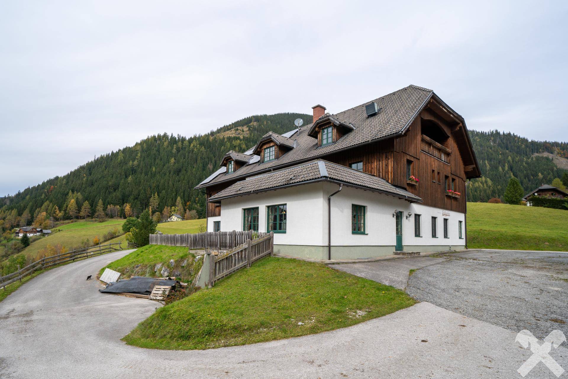 Seitenansicht des Hauptgebäudes mit Holzverkleidung, weißen Wänden und Blick auf die umliegende Berglandschaft und grüne Hügel.