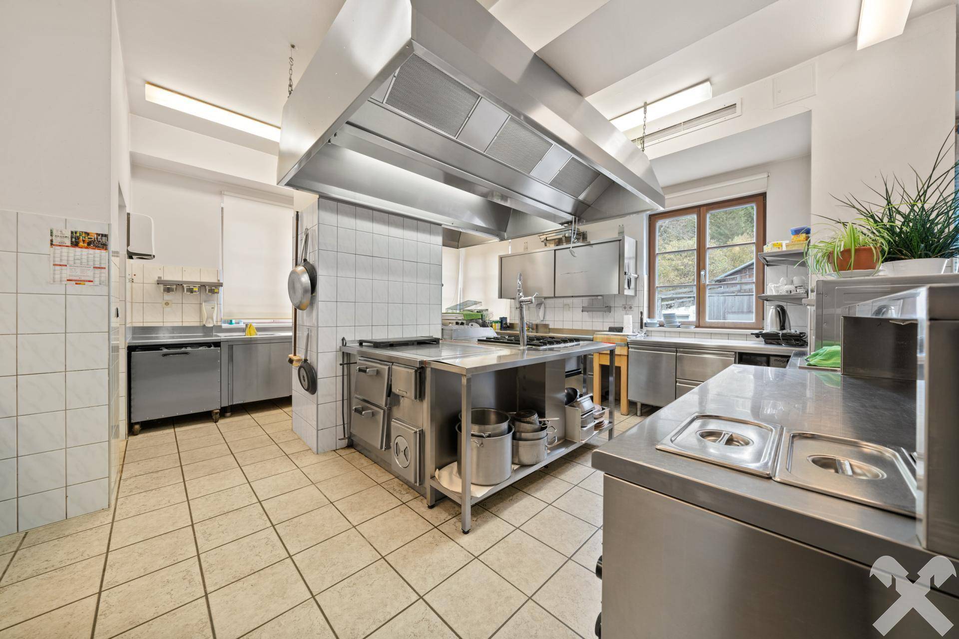 Große, professionelle Küche mit Edelstahloberflächen, Fliesenboden und einer leistungsstarken Ablufthaube, ideal für Gastronomie.