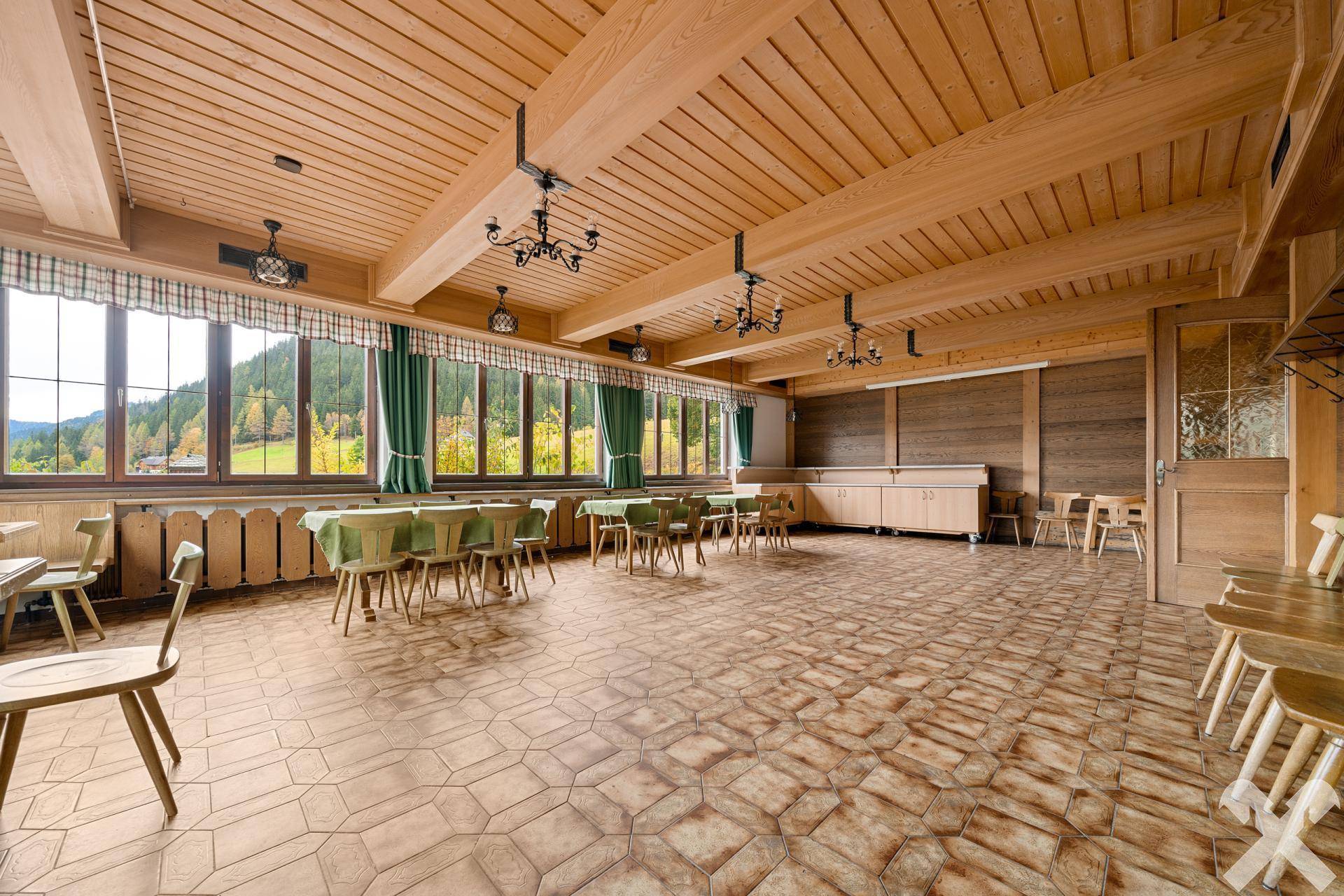 Geräumiger Veranstaltungsraum mit Holzbalkendecke, großen Fenstern und rustikalen Holztischen und Stühlen, ideal für Feiern.