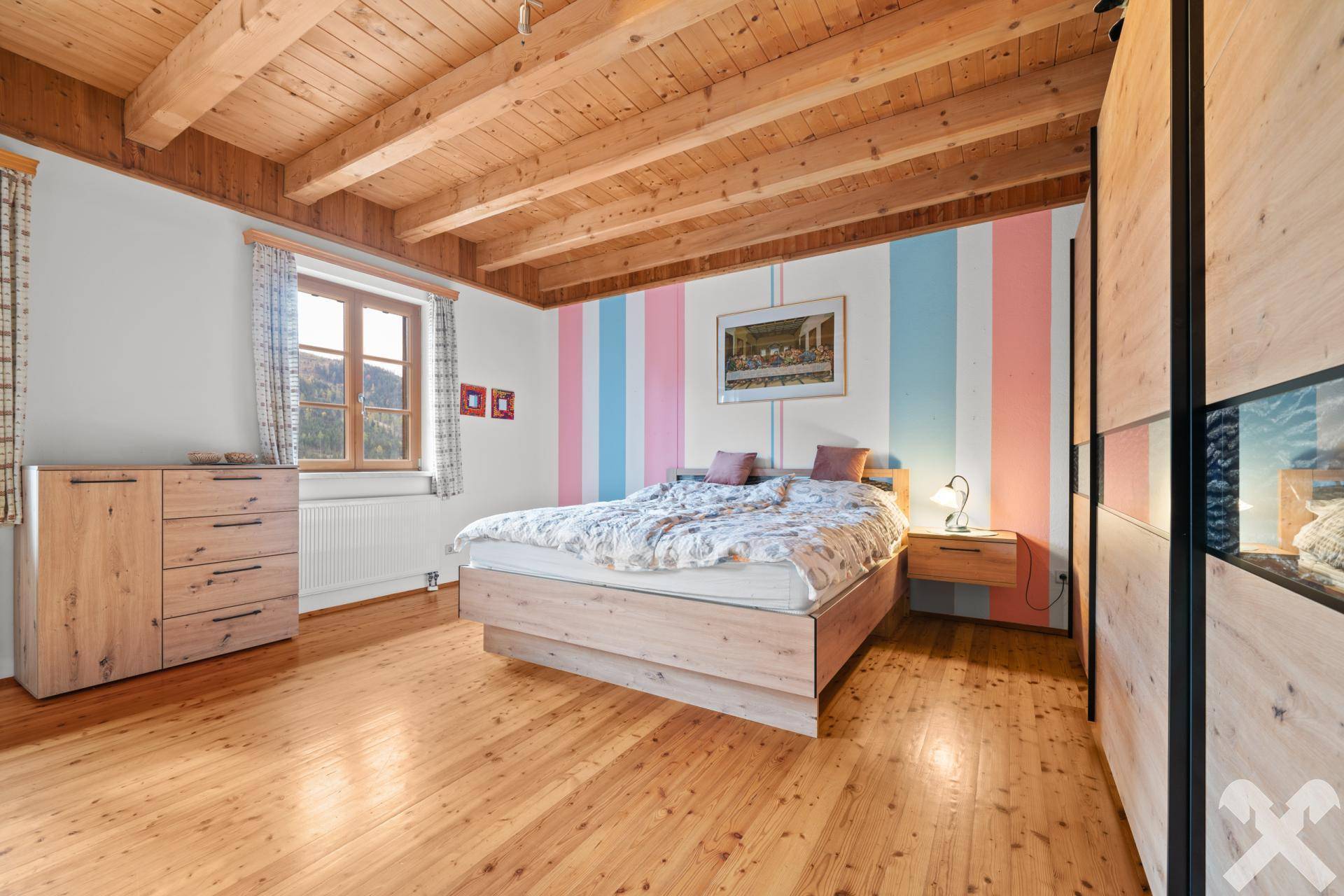 Freundliches Schlafzimmer mit Holzbalkendecke, Holzboden und einer farbenfrohen gestreiften Wand, hell und einladend.