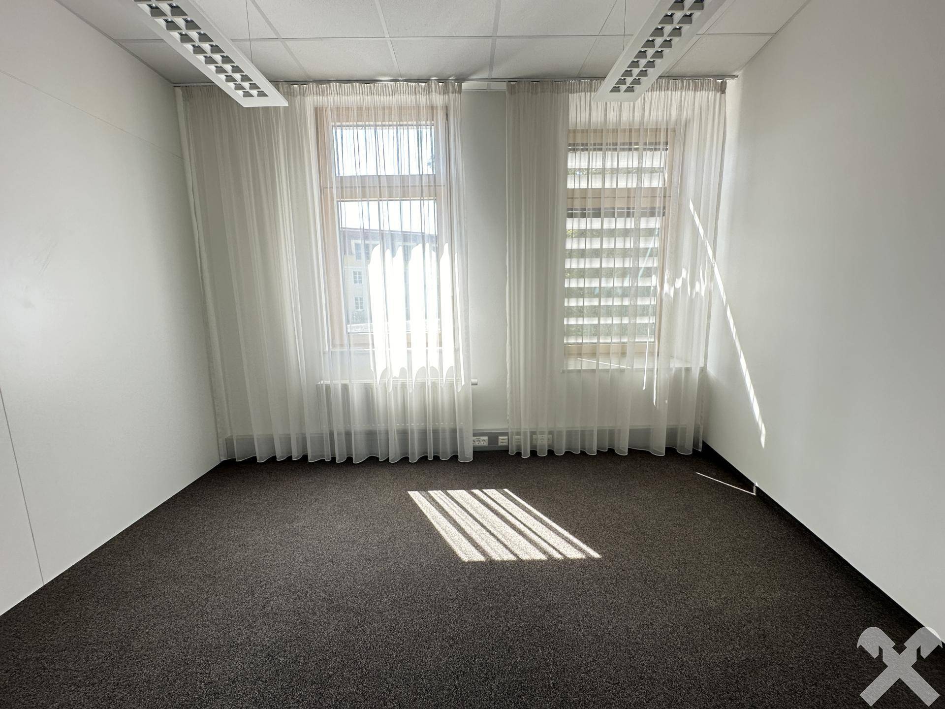 Kleineres Büro mit dunklem Teppichboden, zwei Fenstern mit Vorhängen und Jalousien, bietet angenehmes Tageslicht.