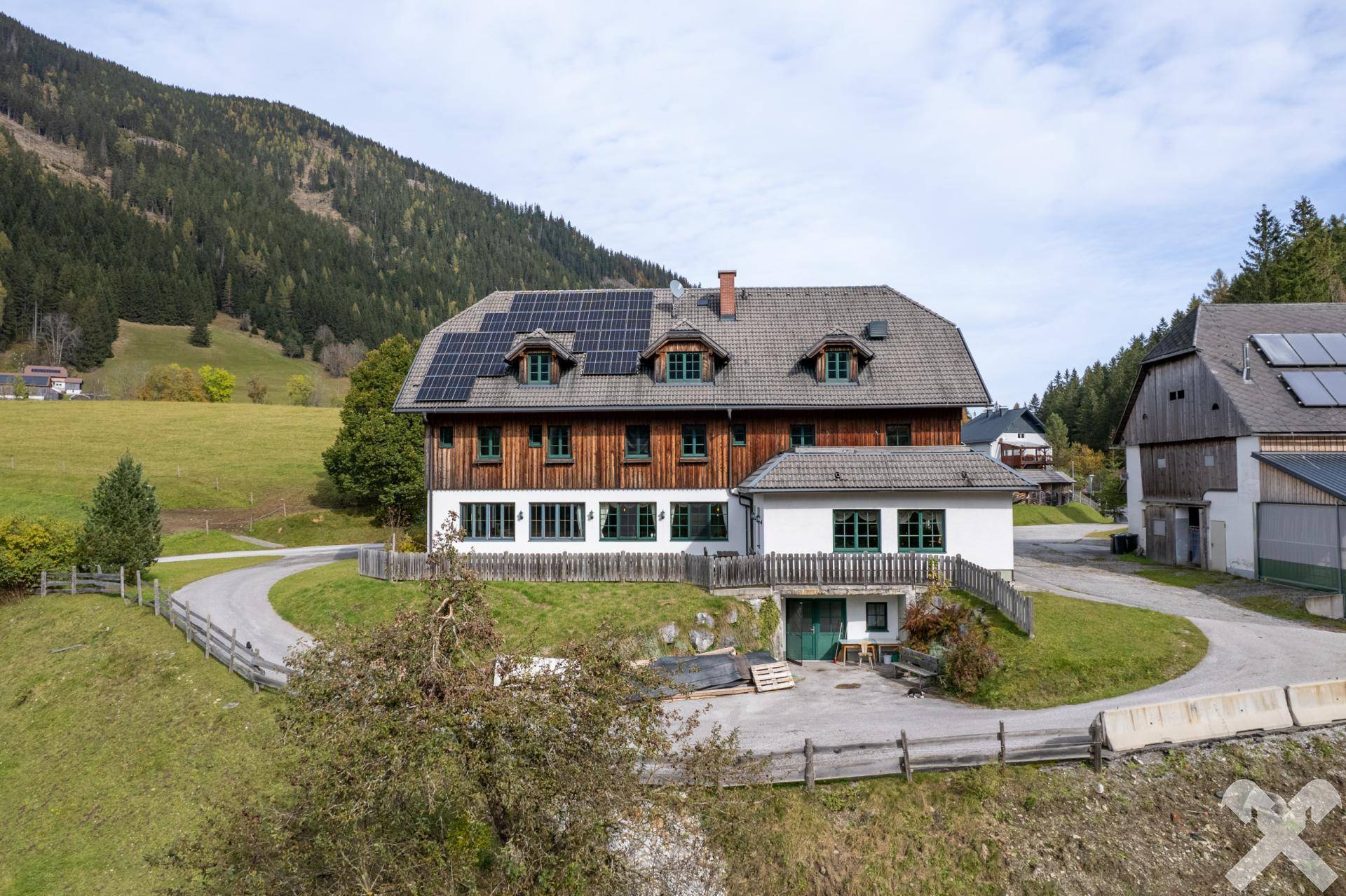 Hauptgebäude mit Holzfassade, weißen Wänden und Solarpaneelen auf dem Dach, eingebettet in eine ländliche Bergkulisse.