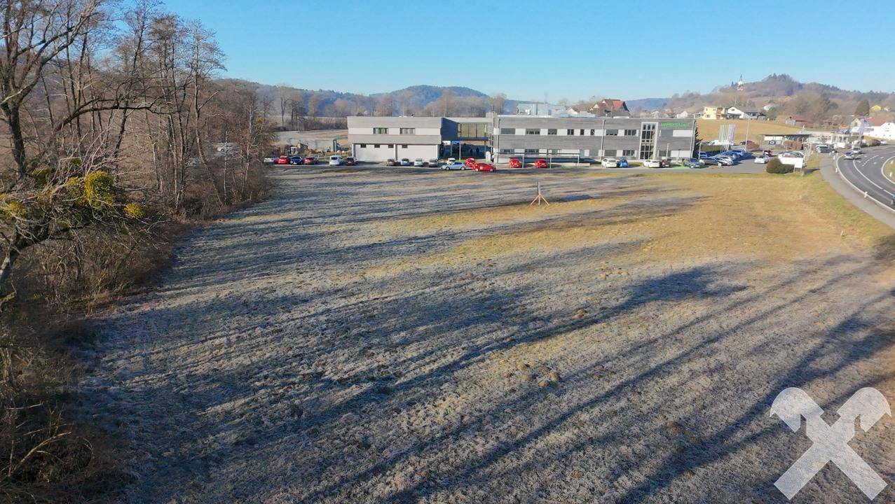 Luftaufnahme eines weitläufigen Grundstücks neben einer Straße mit Gewerbegebäuden und Hügeln im Hintergrund.