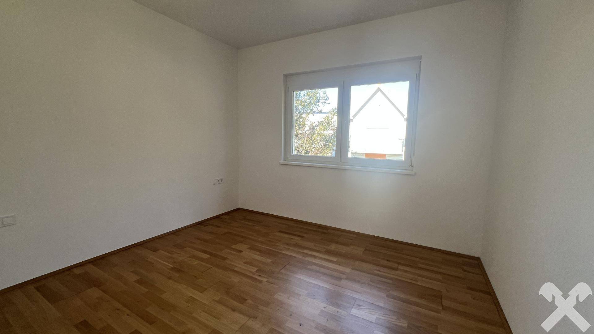 Leeres Zimmer mit Holzboden und einem Fenster, das viel Tageslicht hereinlässt.