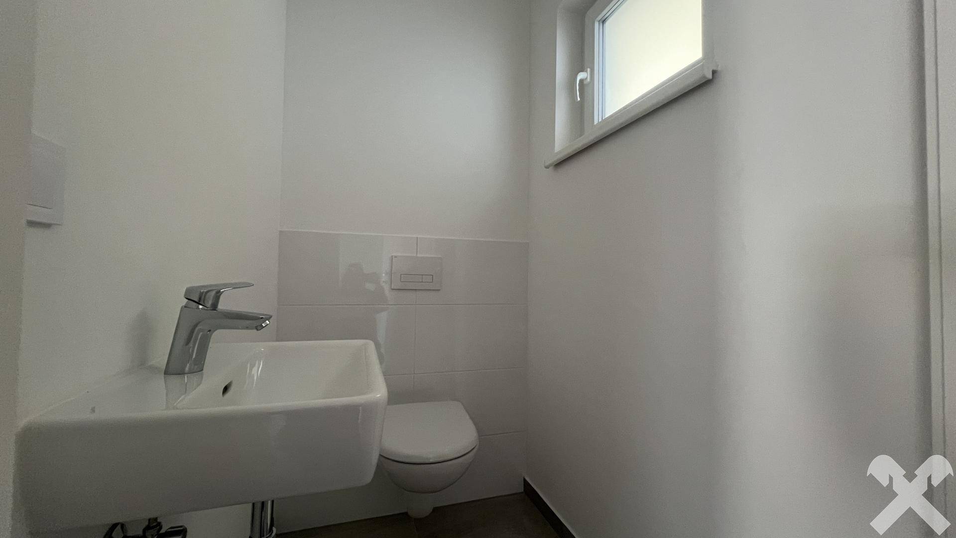 Separates WC mit wandhängender Toilette, kleinem Waschbecken und Fenster.