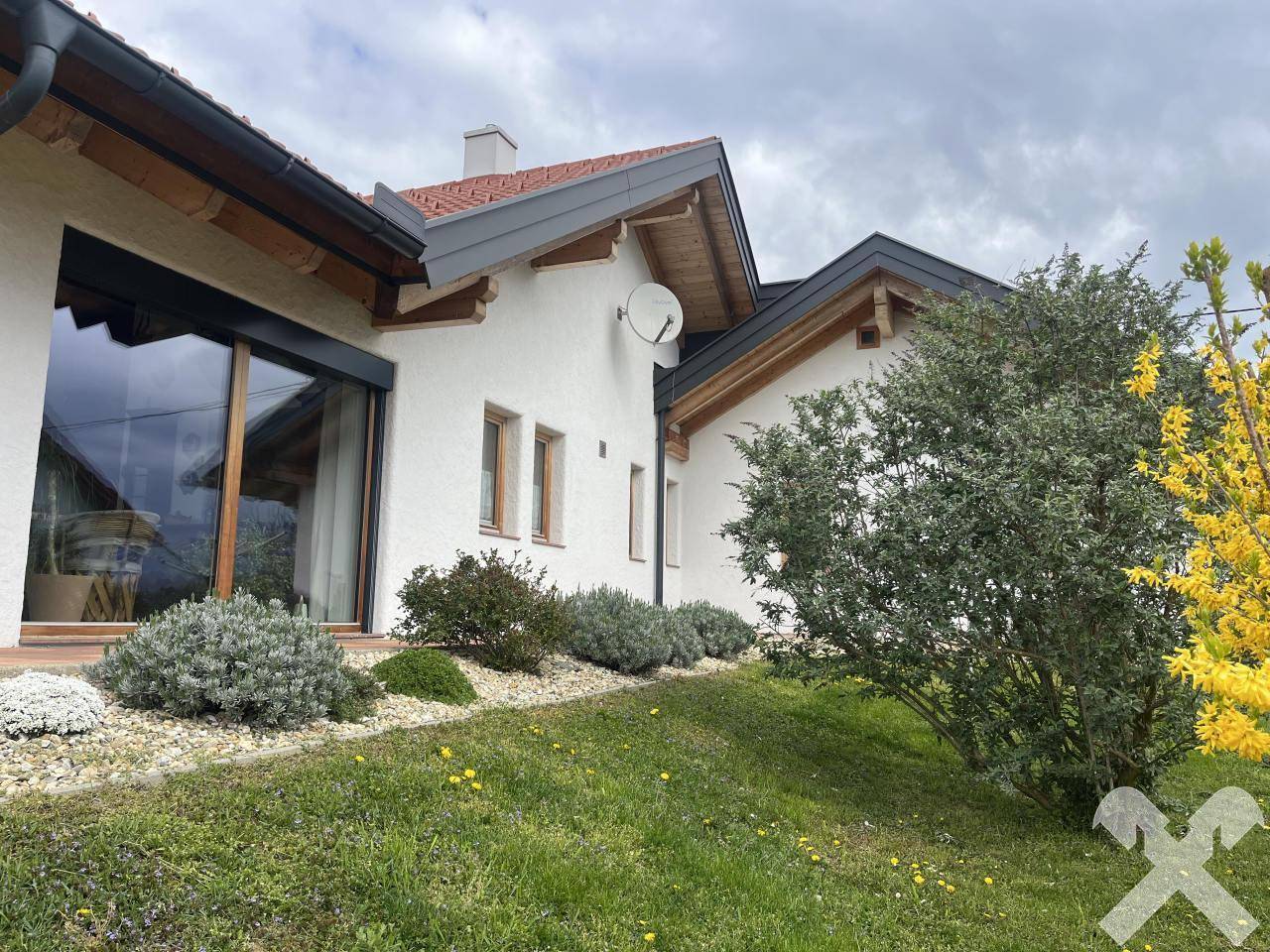 Seitenansicht des Hauses mit großen Fenstern, Holzdetails und blühenden Sträuchern.