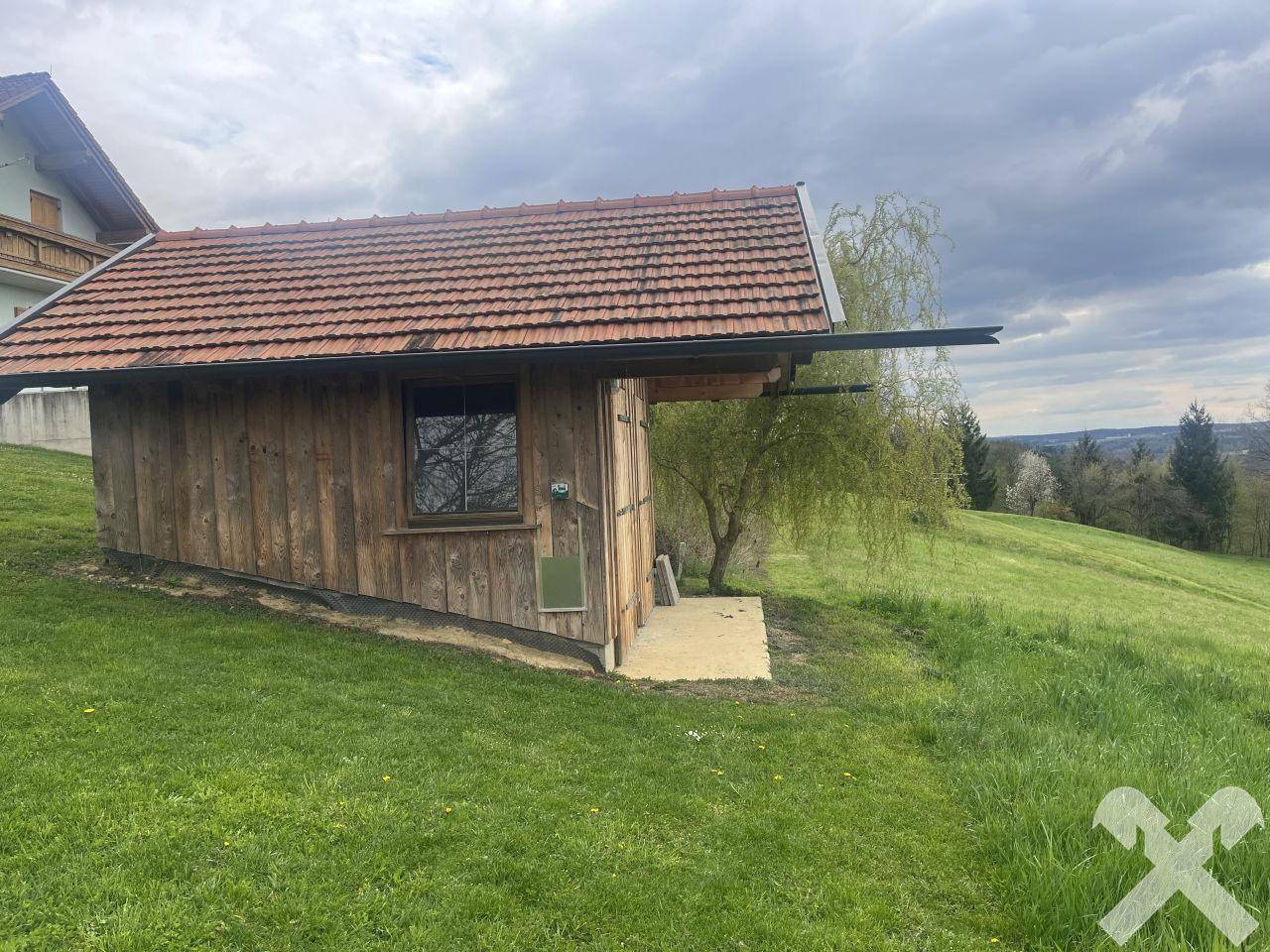 Rustikales Gartenhaus aus Holz mit Ziegeldach, eingebettet in die grüne Landschaft.
