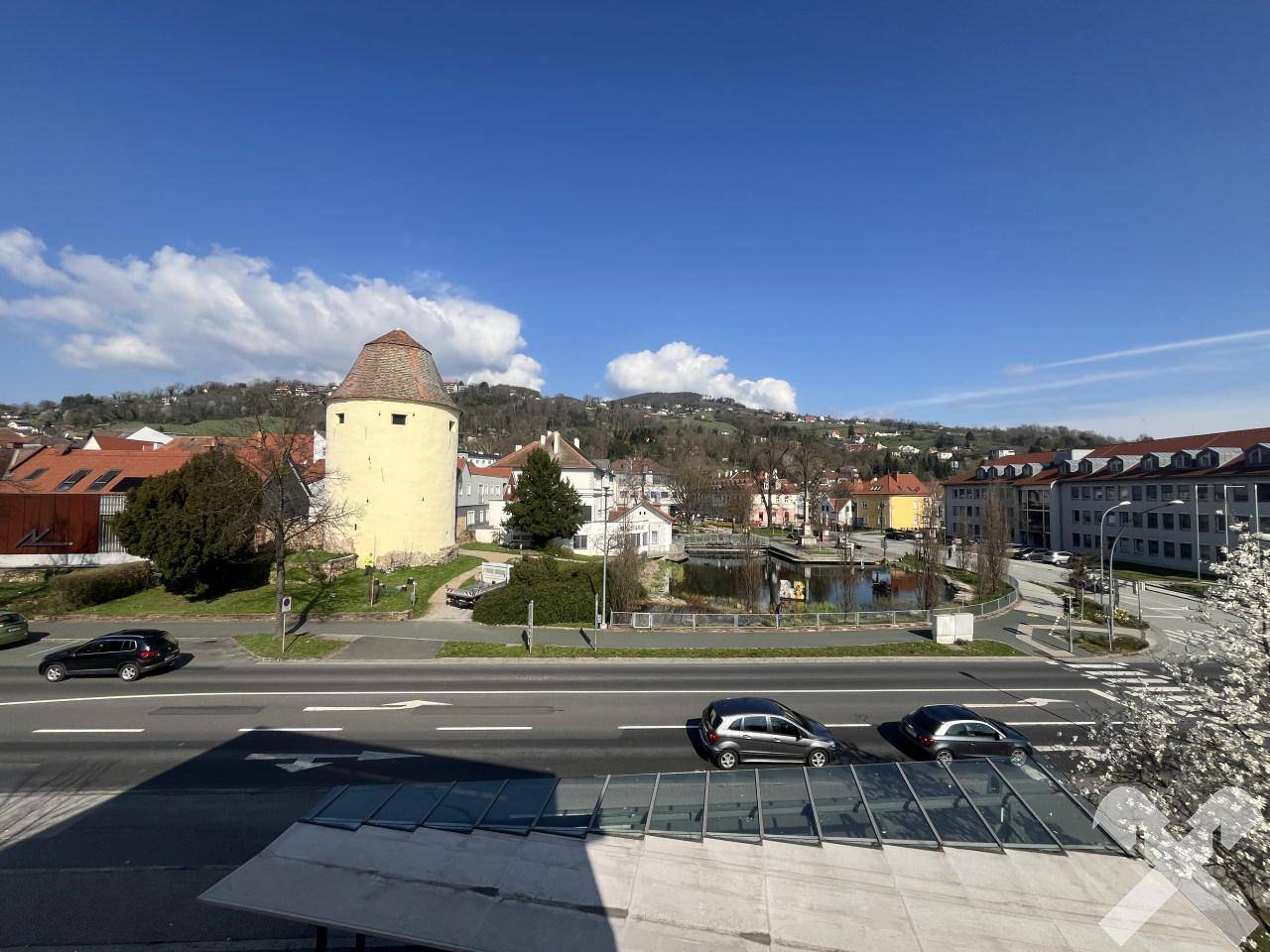 Weitblick von einem Fenster auf eine Straße mit Autos, einen historischen Turm und grüne Hügel im Hintergrund.