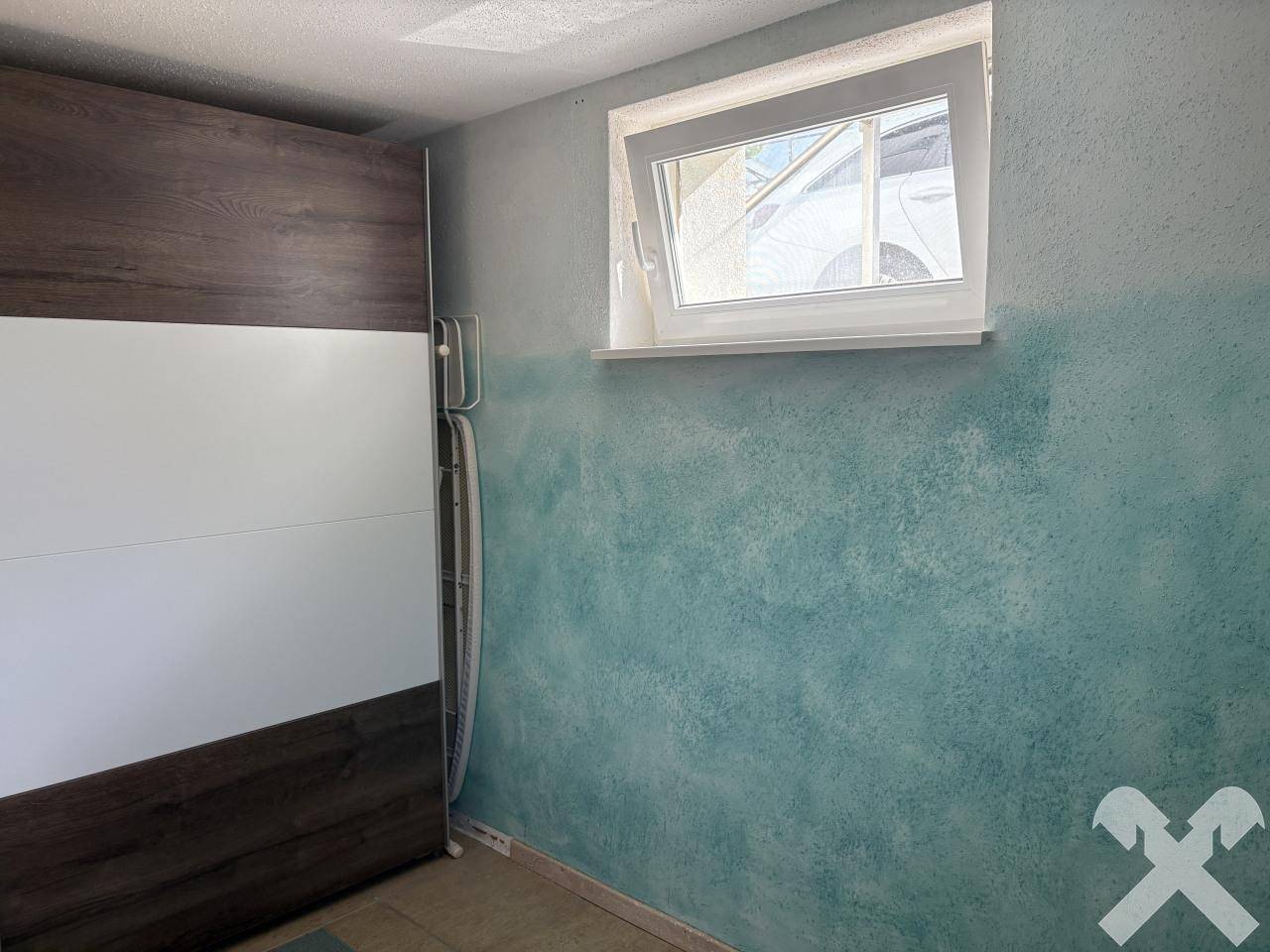 Raum mit Kleiderschrank und einem Fenster, die Wand ist zweifarbig in Blaugrün und Weiß gestrichen.
