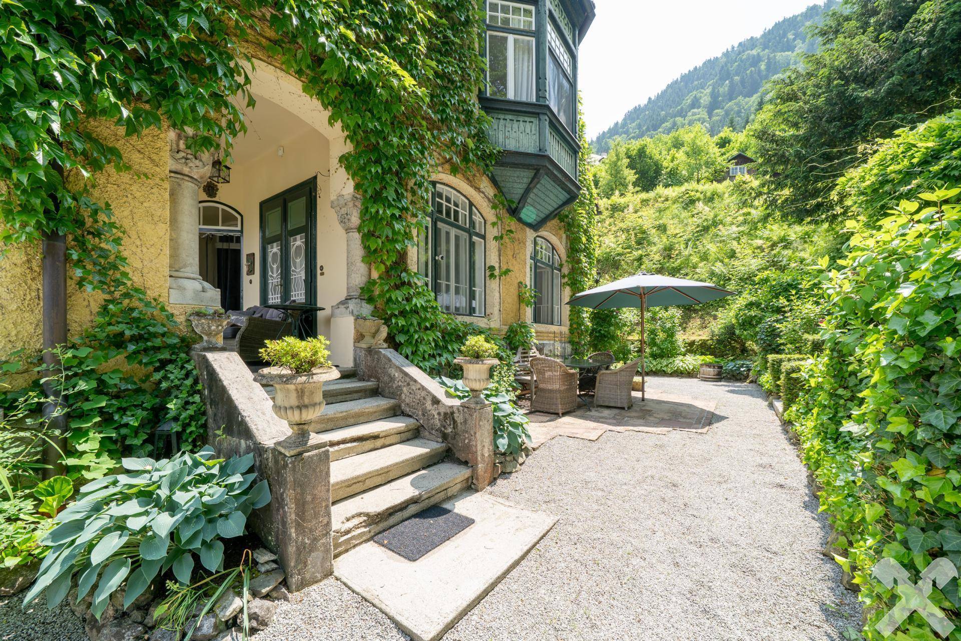 Eingangsbereich der Villa mit Steintreppe und kleiner Terrasse mit Sitzgelegenheiten im Grünen.