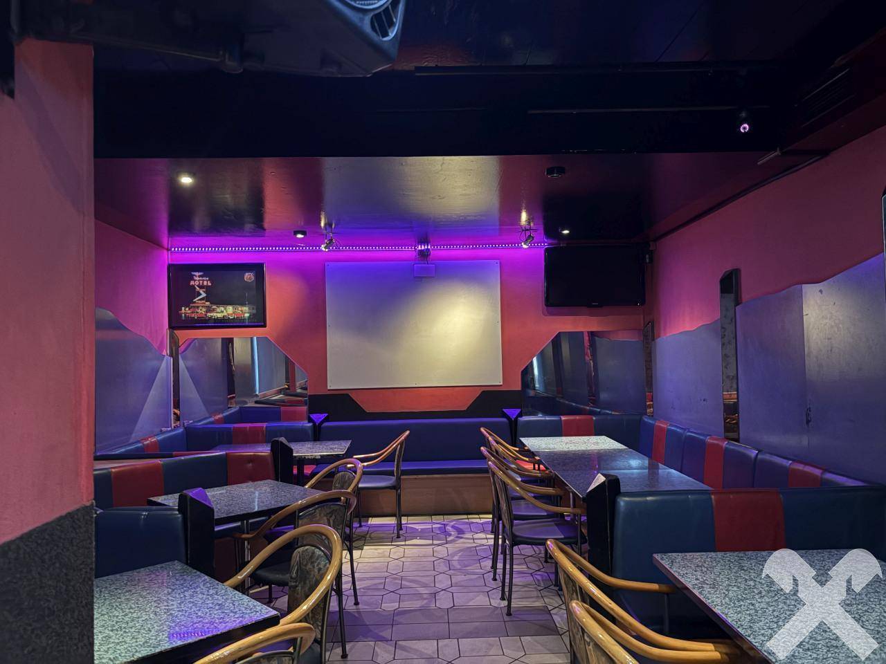 Bar- oder Clubbereich mit gepolsterten Sitzbänken, Tischen und atmosphärischer violetter Beleuchtung.