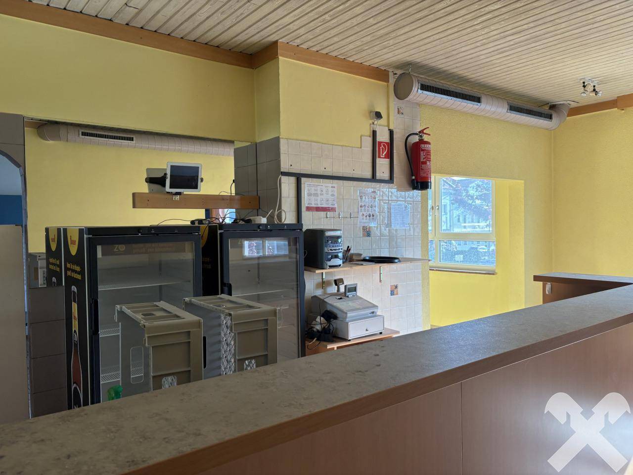 Gastronomiebereich mit Theke, Kühlvitrinen und Lüftungsrohren unter einer Holzdecke.