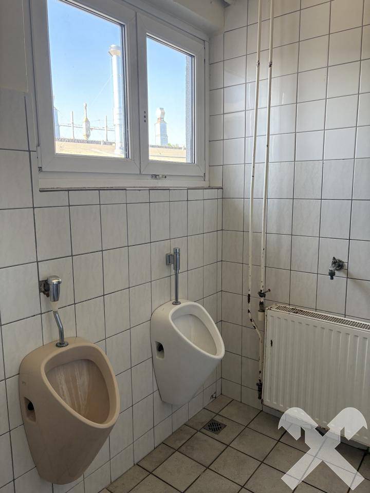 Herren-WC mit zwei Urinalen, gefliesten Wänden und einem Fenster für Tageslicht.