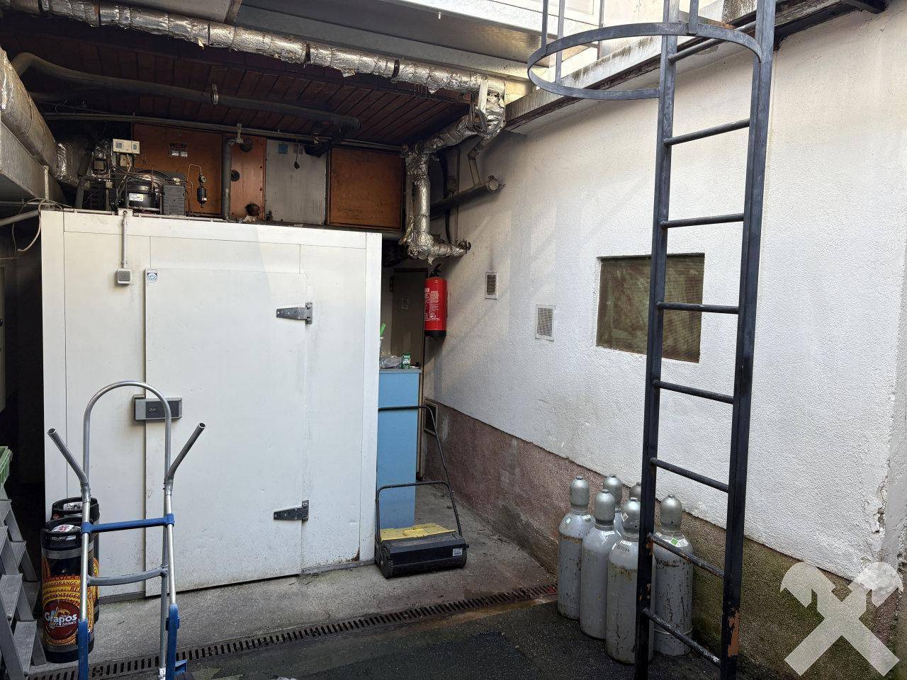 Außenbereich mit industriellen Anlagen, Rohren und einem Kühlraum an der Gebäudewand.