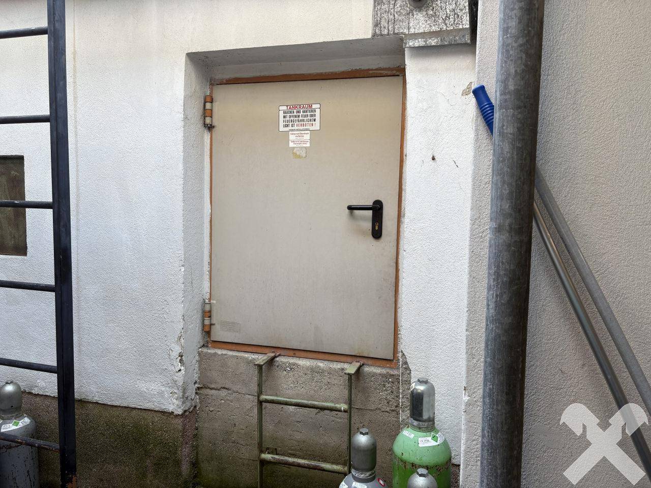 Eine einfache Metalltür in einer weißen Wand, umgeben von industriellen Elementen und Leitern.