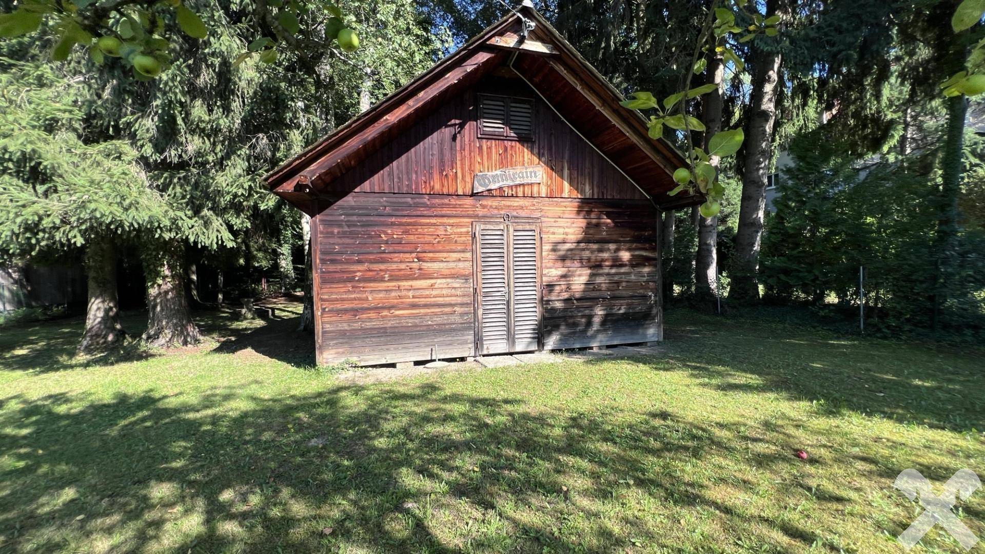 Detailansicht eines rustikalen Gartenhäuschens aus Holz, umgeben von hohen Bäumen.