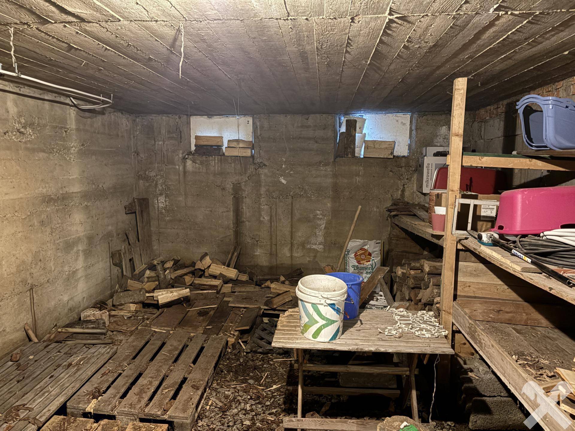 Unausgebauter Kellerraum mit Holzstapeln und Lagerregalen.