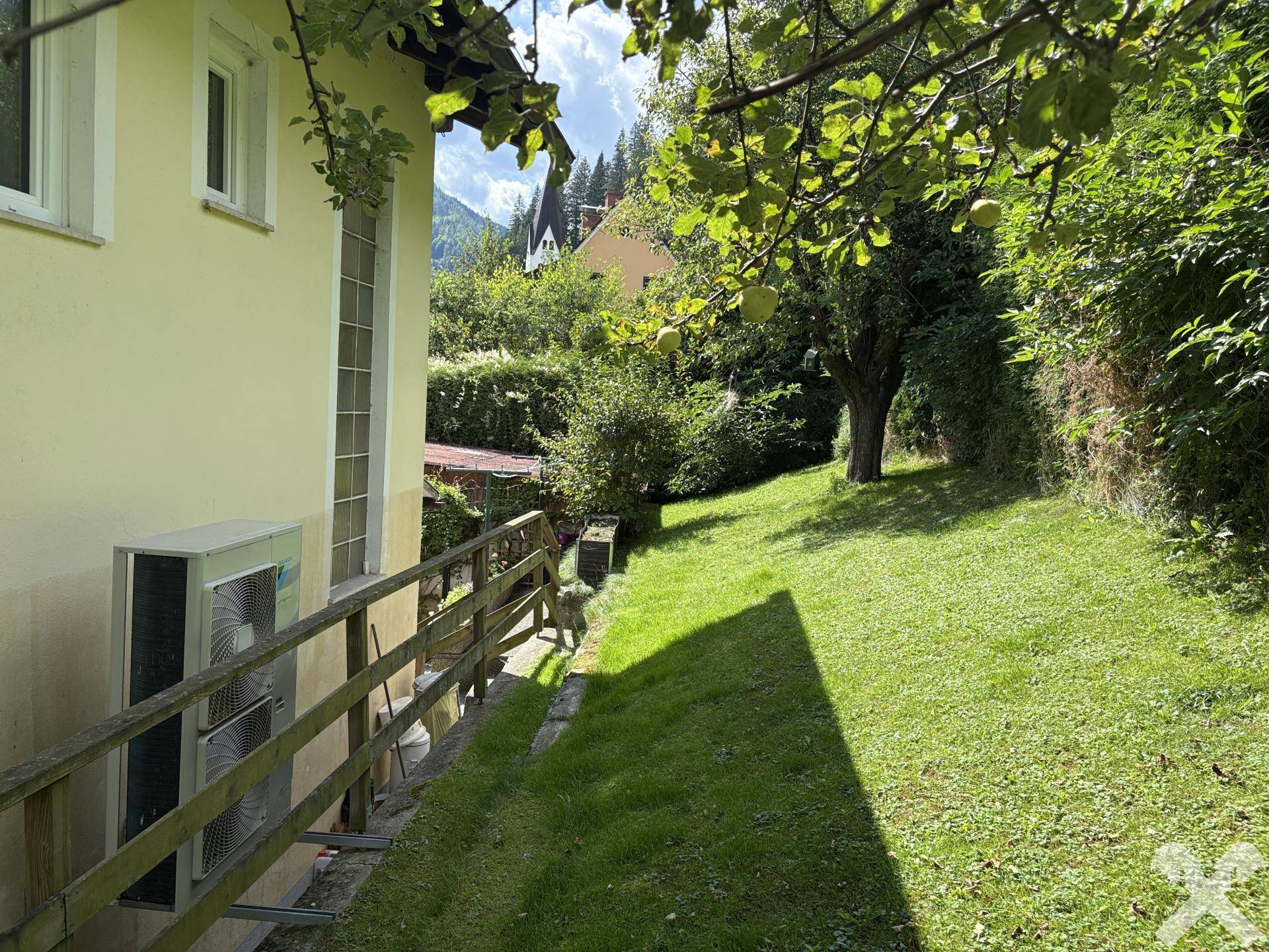 Schmaler Gartenstreifen neben dem Haus mit Rasen, Bäumen und einer Außeneinheit.