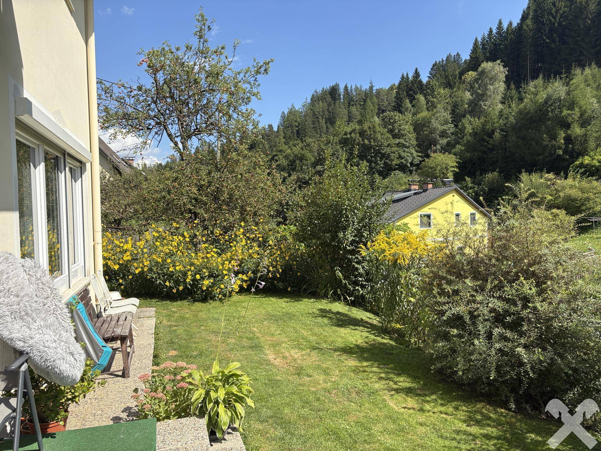 Weitläufiger Gartenbereich mit Rasen, Blumen und einer Holzbank, umgeben von viel Grün.