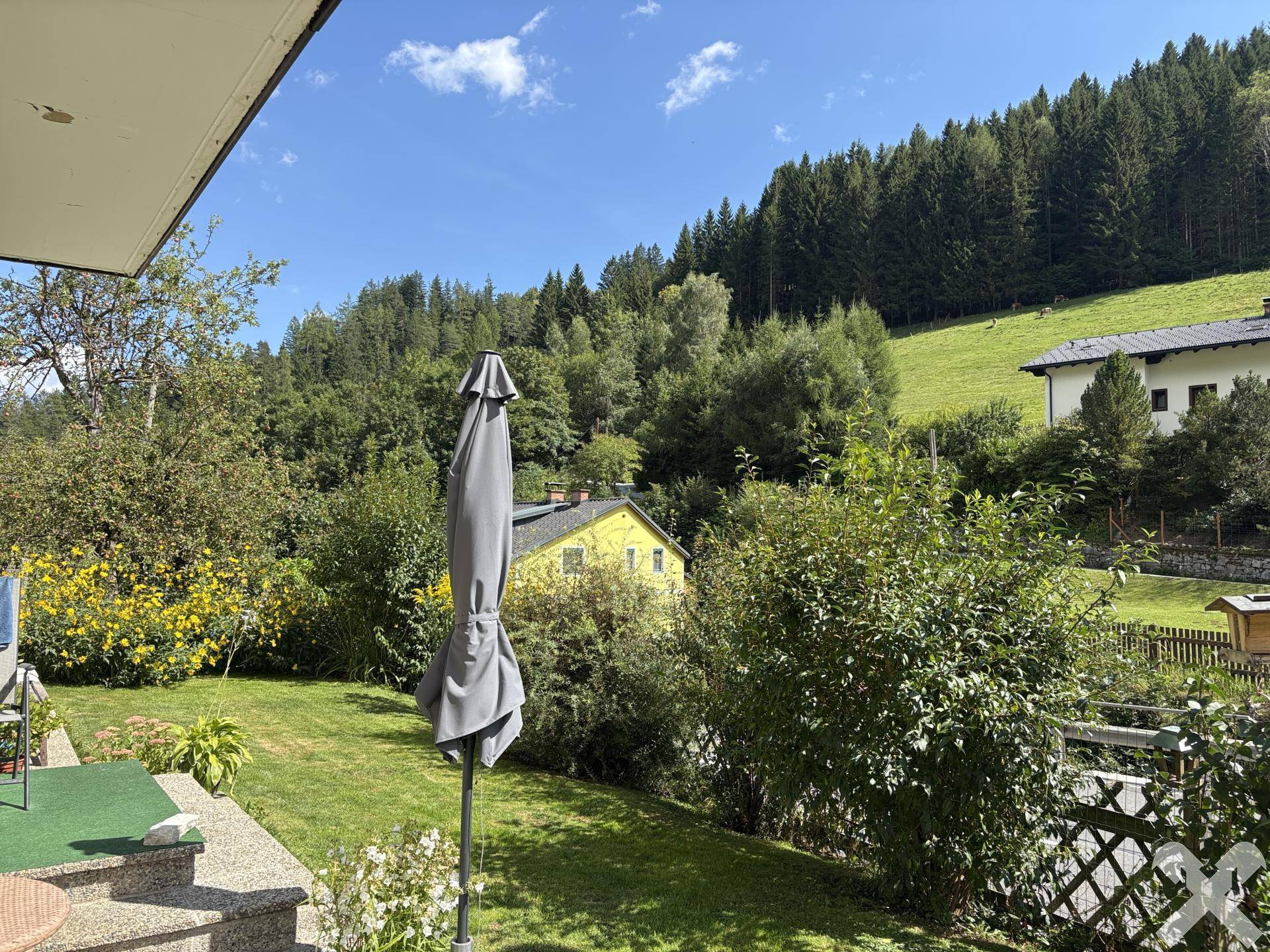 Blick vom Garten auf die umliegende grüne Hügellandschaft und weitere Häuser.