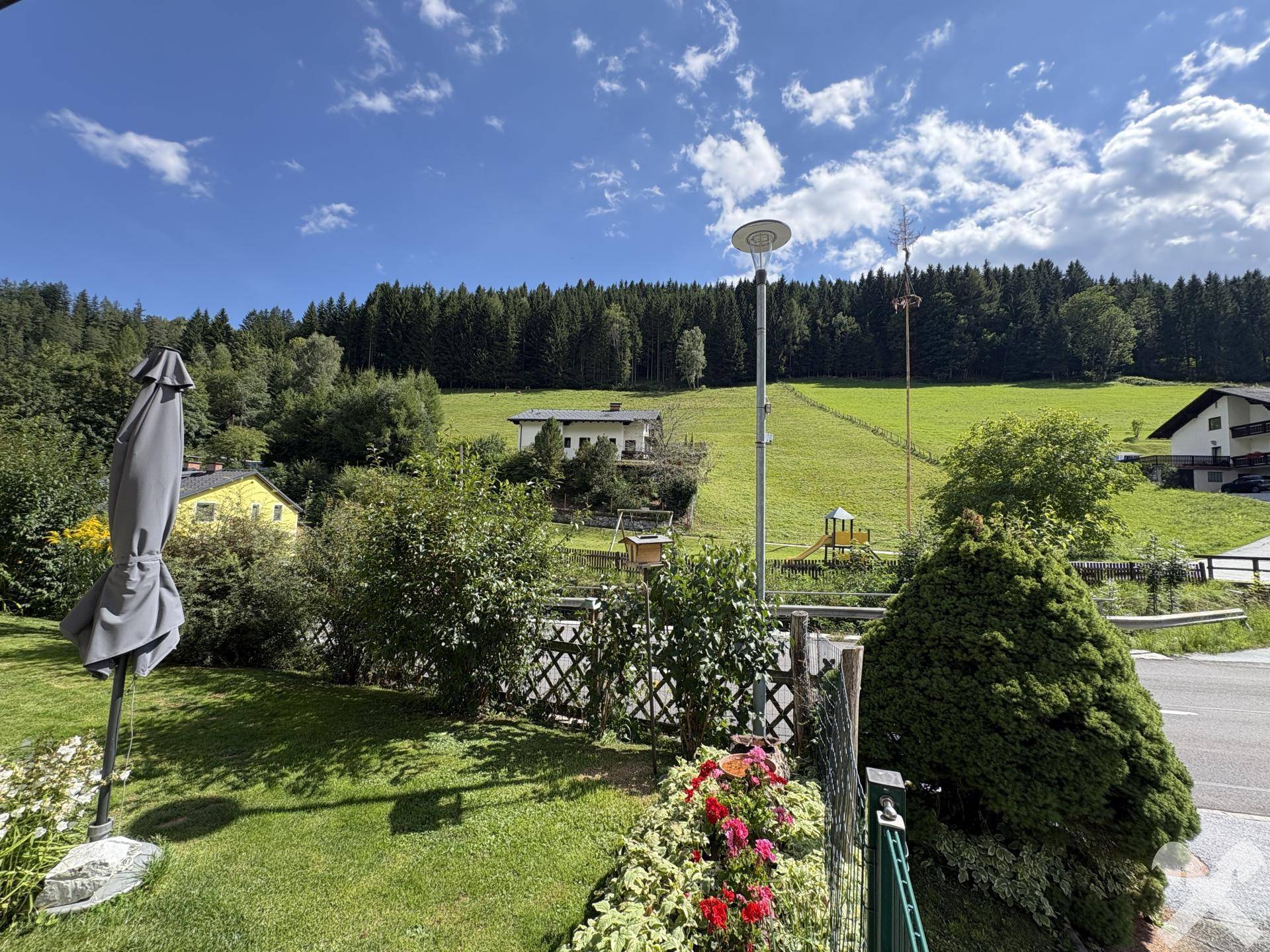 Gartenansicht mit Blick auf die Straße, Blumenbeete und die bewaldeten Hügel.