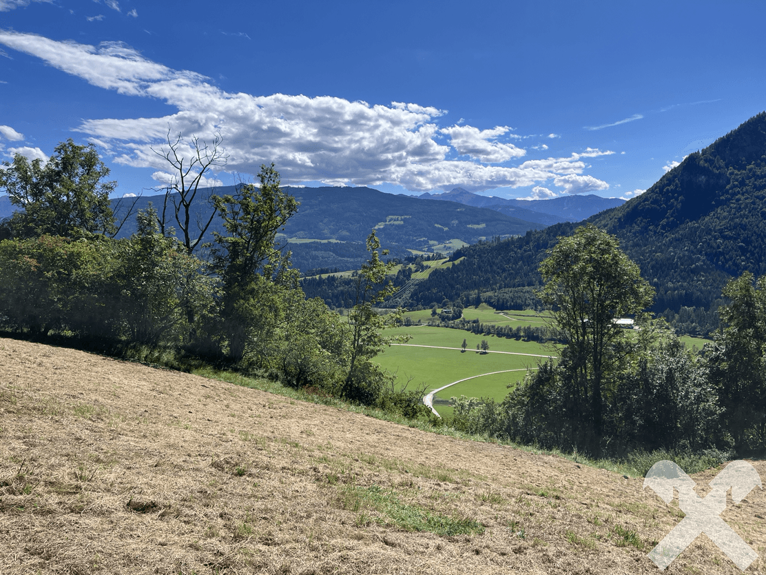 Weitläufige Landschaft mit grünen Feldern, Wäldern und fernen Bergen unter einem blauen Himmel.