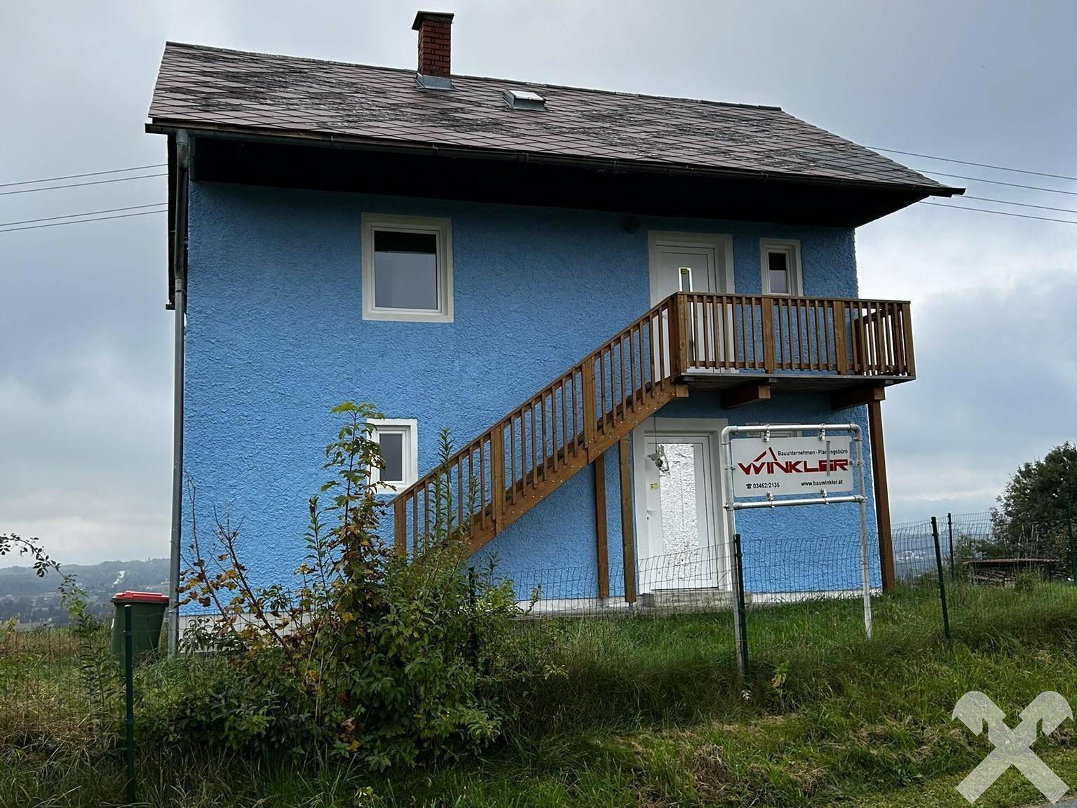 Vorderansicht des blauen Hauses mit Außentreppe und einem Verkaufsschild.