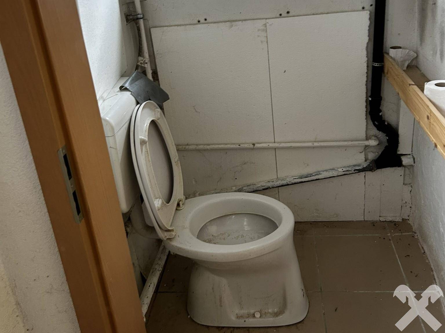 Einfache Toilette in einem Raum mit sichtbaren Rohren und Fliesenboden, der renovierungsbedürftig ist.