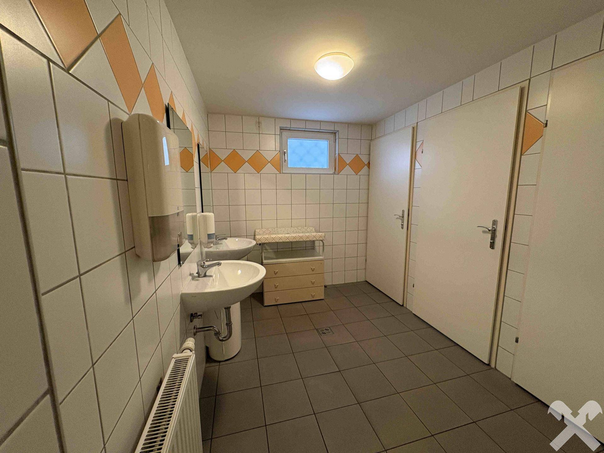 Öffentliche Toilettenanlage mit zwei Waschbecken, Fliesenboden und einem Wickeltisch.