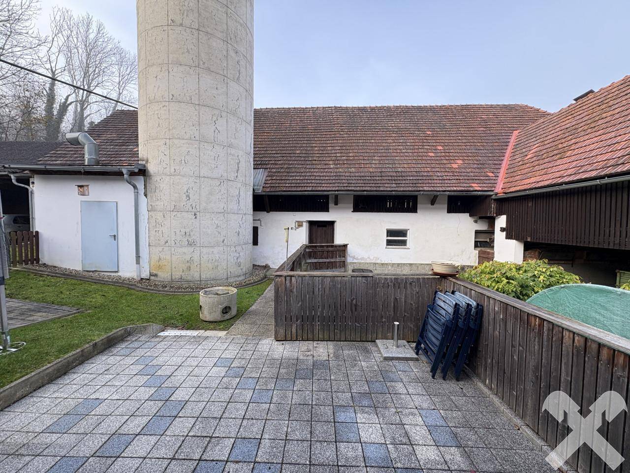 Gepflasterte Terrasse neben einem hohen Silo und einem Nebengebäude, mit Rasenfläche und Holzzaun.