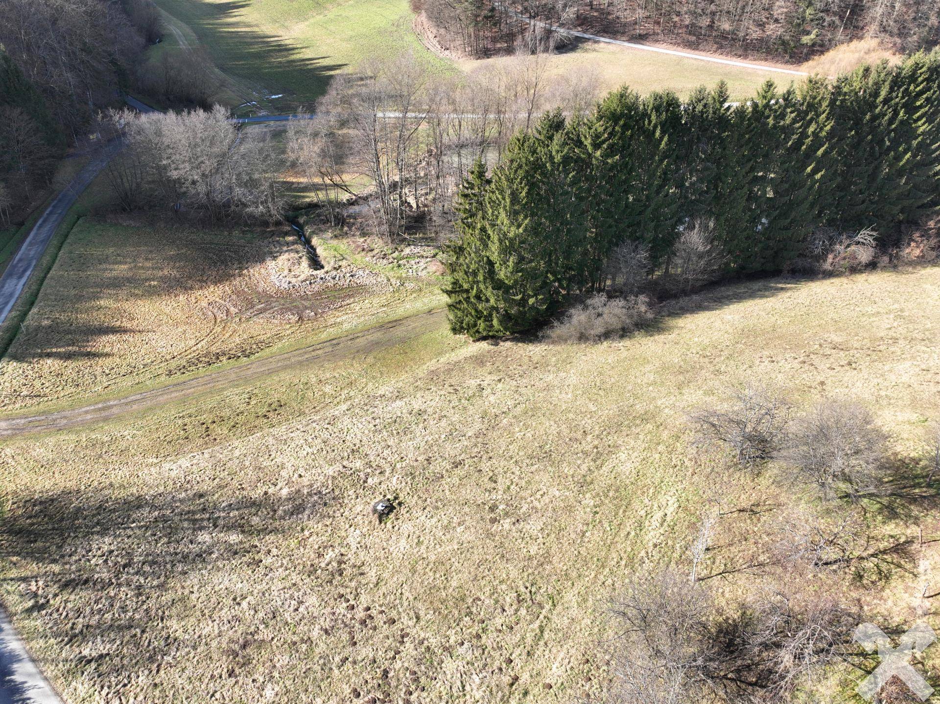 Weitläufige Luftaufnahme eines Grundstücks mit Feldern, Waldrand und einem kleinen Gewässer in hügeliger Landschaft.