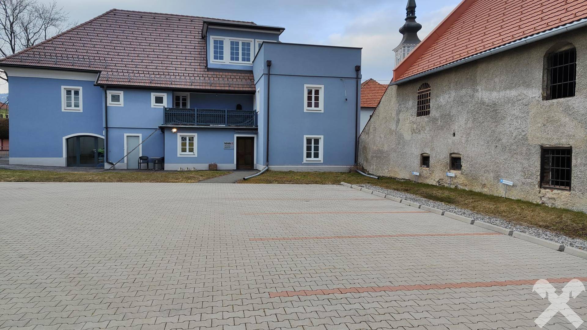 Ansicht des blauen Gebäudes von einem gepflasterten Parkplatz aus, mit einem Kirchturm im Hintergrund.