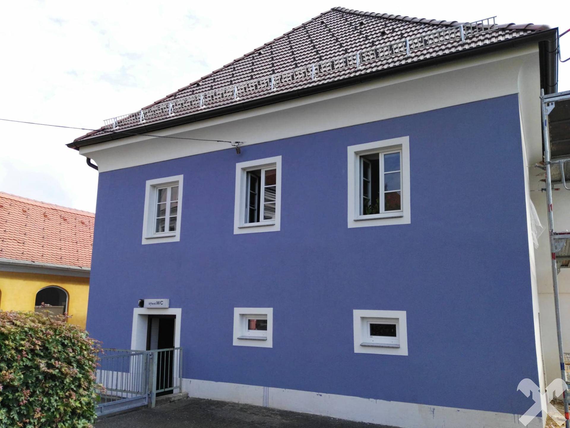 Ansicht des blauen Gebäudes mit Rampe und Satteldach, umgeben von einer Hecke und anderen Häusern.